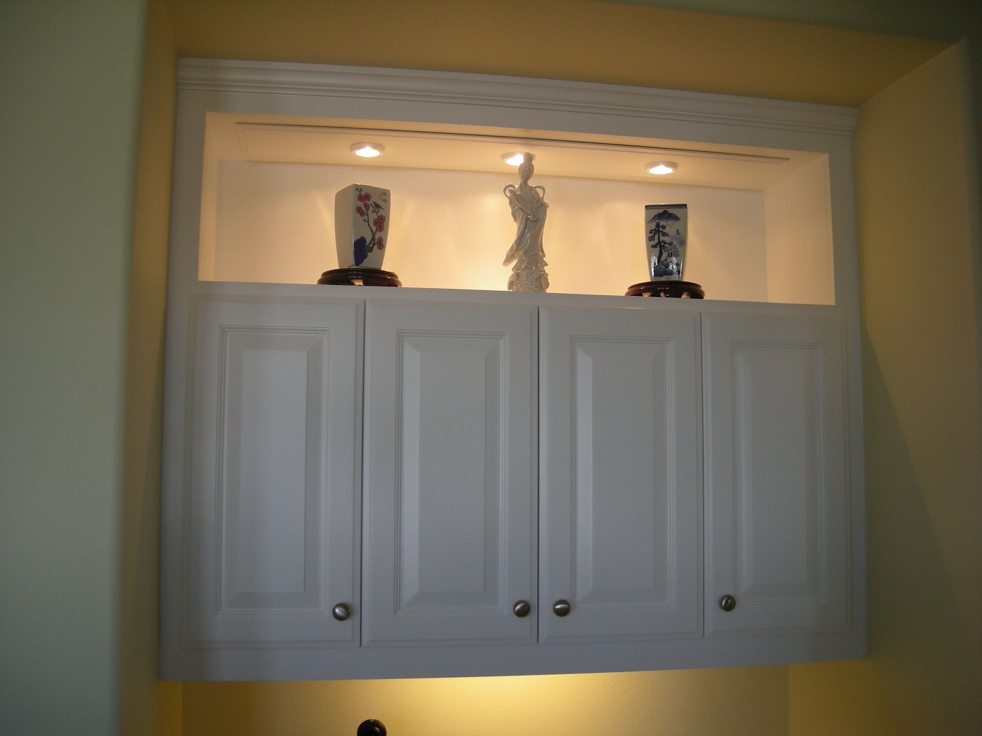 White cabinet with display niche above, lit by recessed lights, holding porcelain vases.