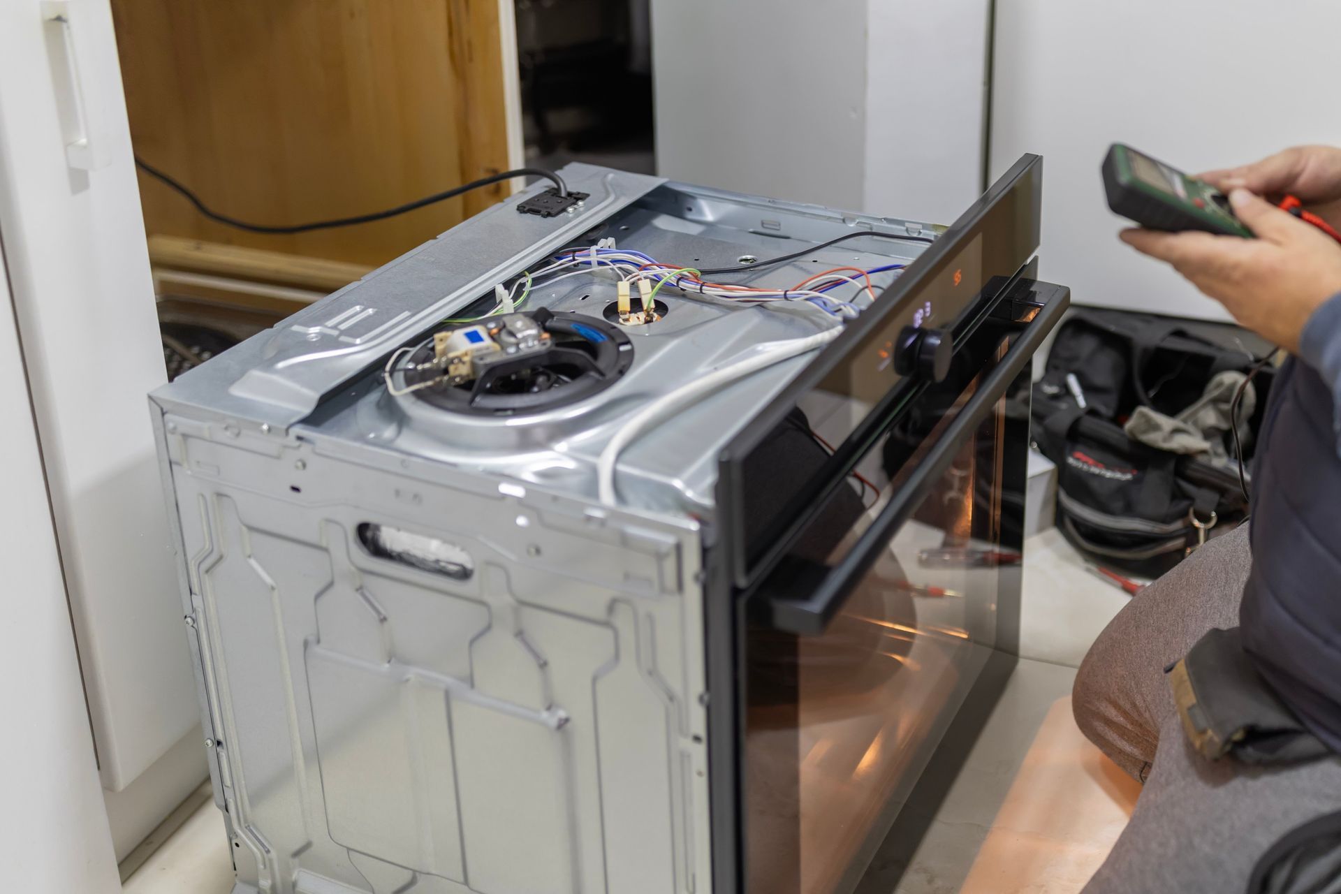 Person repairing a stainless steel oven with the top removed, using a remote control in a kitchen.