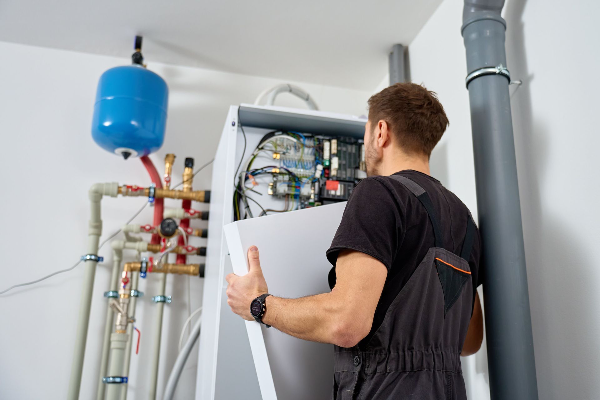 Technician inspecting a wall-mounted boiler with exposed pipes and a blue expansion tank in a utility room