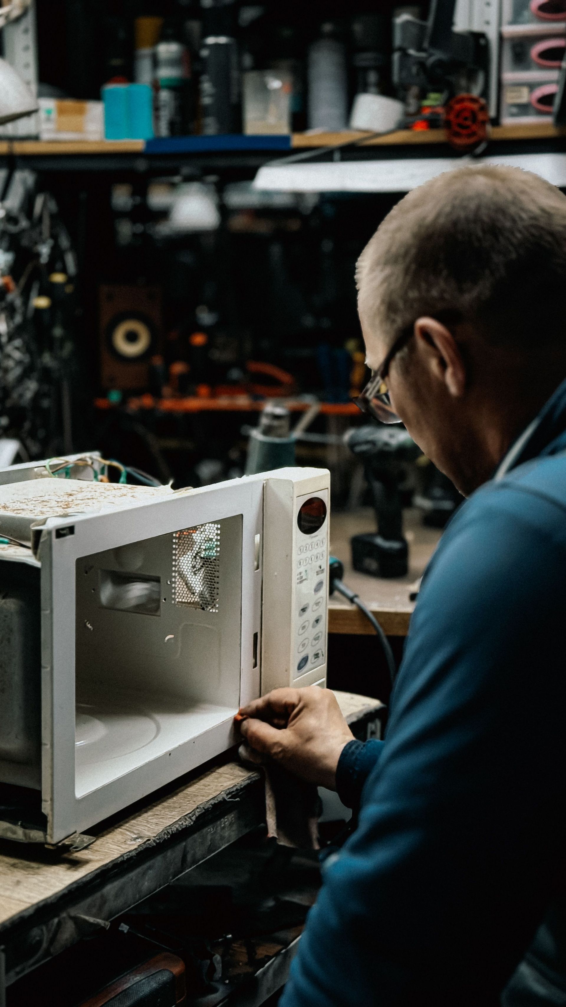Person repairing a white microwave in a cluttered workshop, viewed from behind
