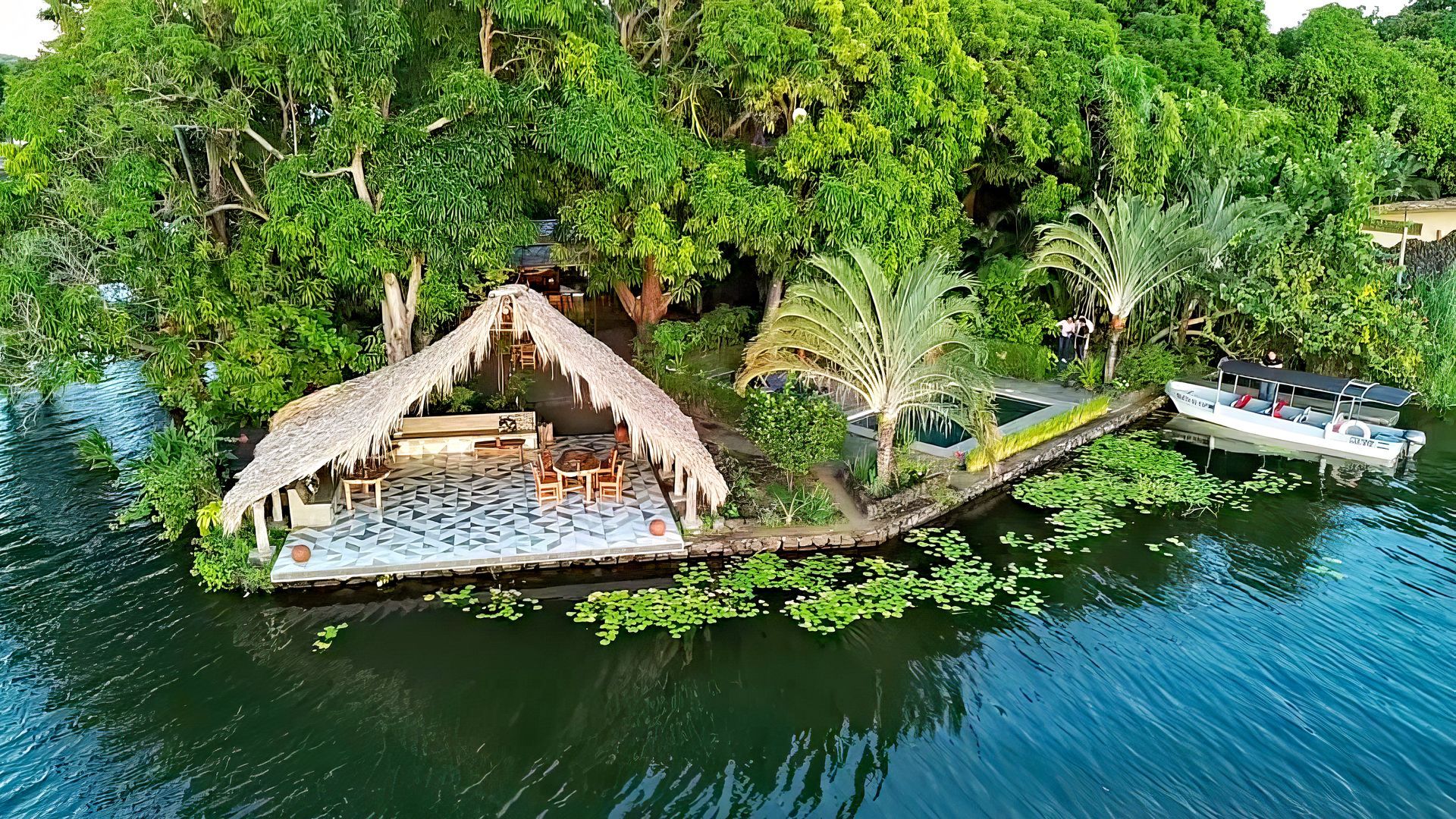 Island with a thatched-roof structure, boat, and lush greenery in a body of water.