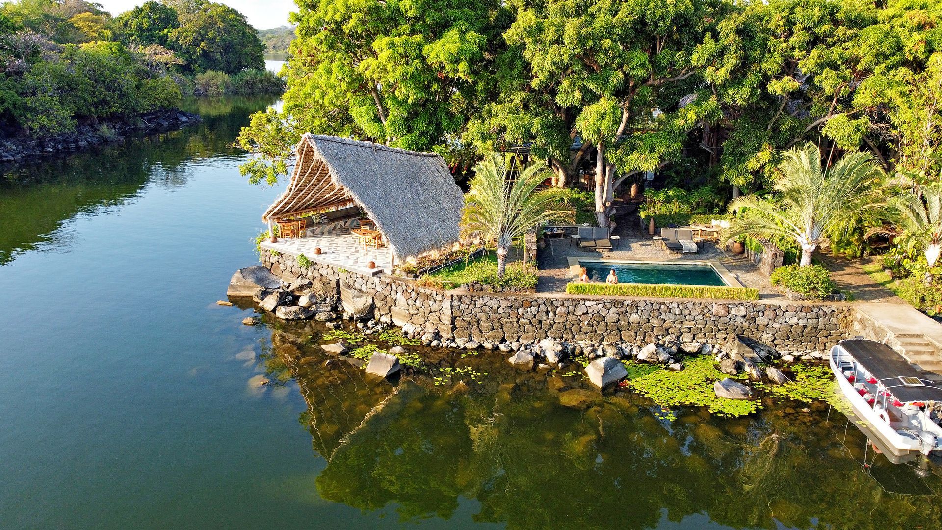 A waterside thatched-roof structure, stone walls, and a pool surrounded by lush trees.