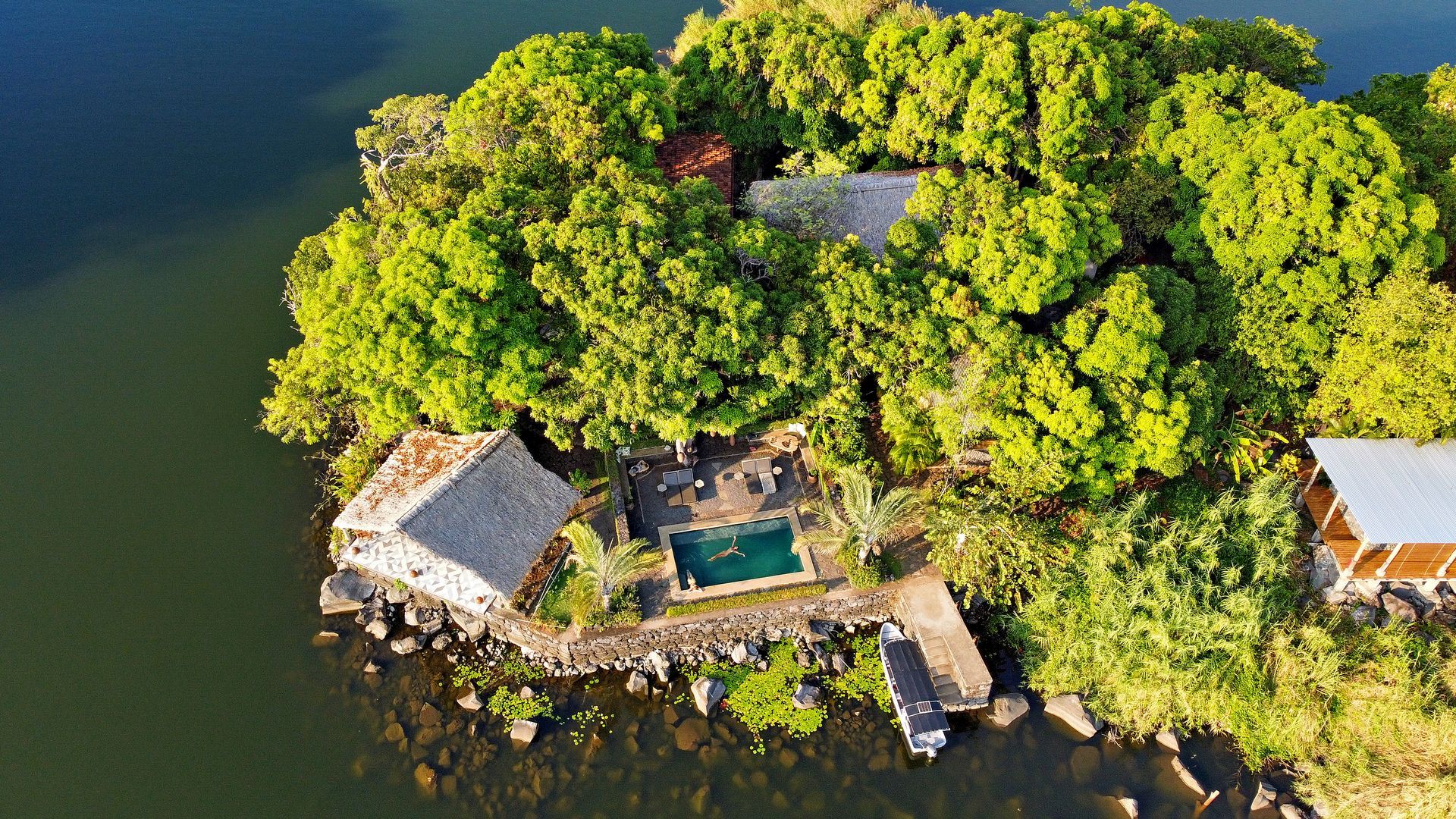Small island with lush green trees and buildings.  A swimming pool, dock and boat are visible.