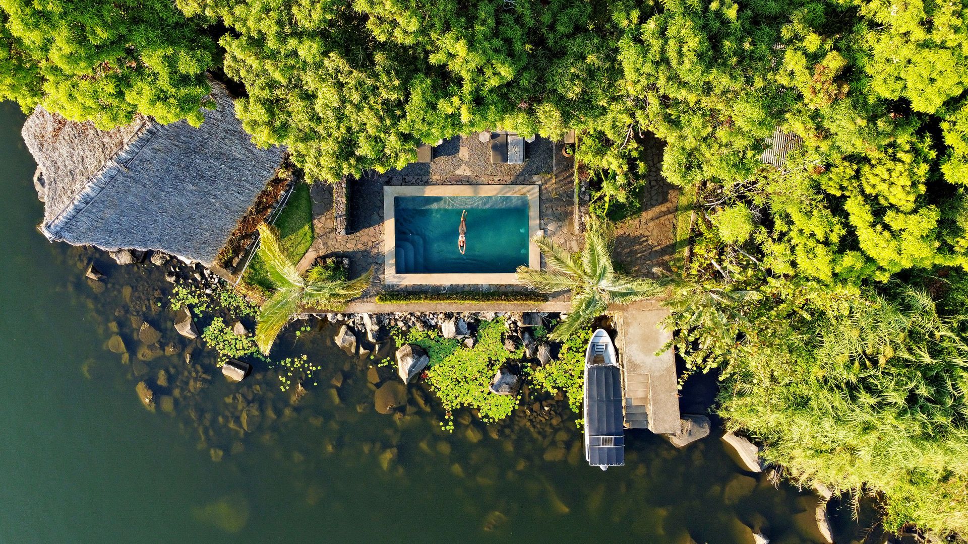 Aerial view of a stone pool and thatched roof building on an island, surrounded by lush greenery and water.