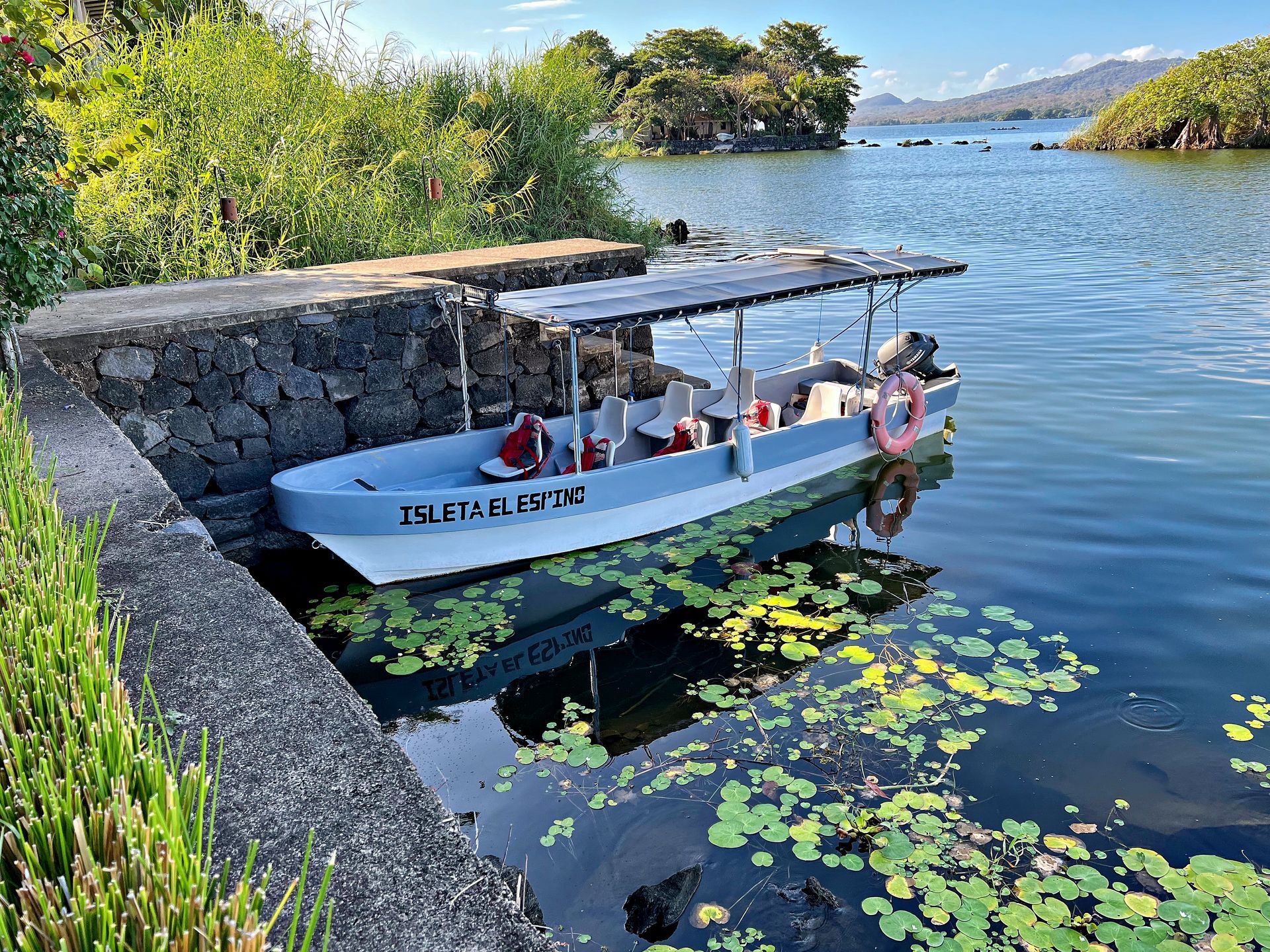 Boat docked at stone pier on a lake, lily pads, lush greenery, island in the distance.