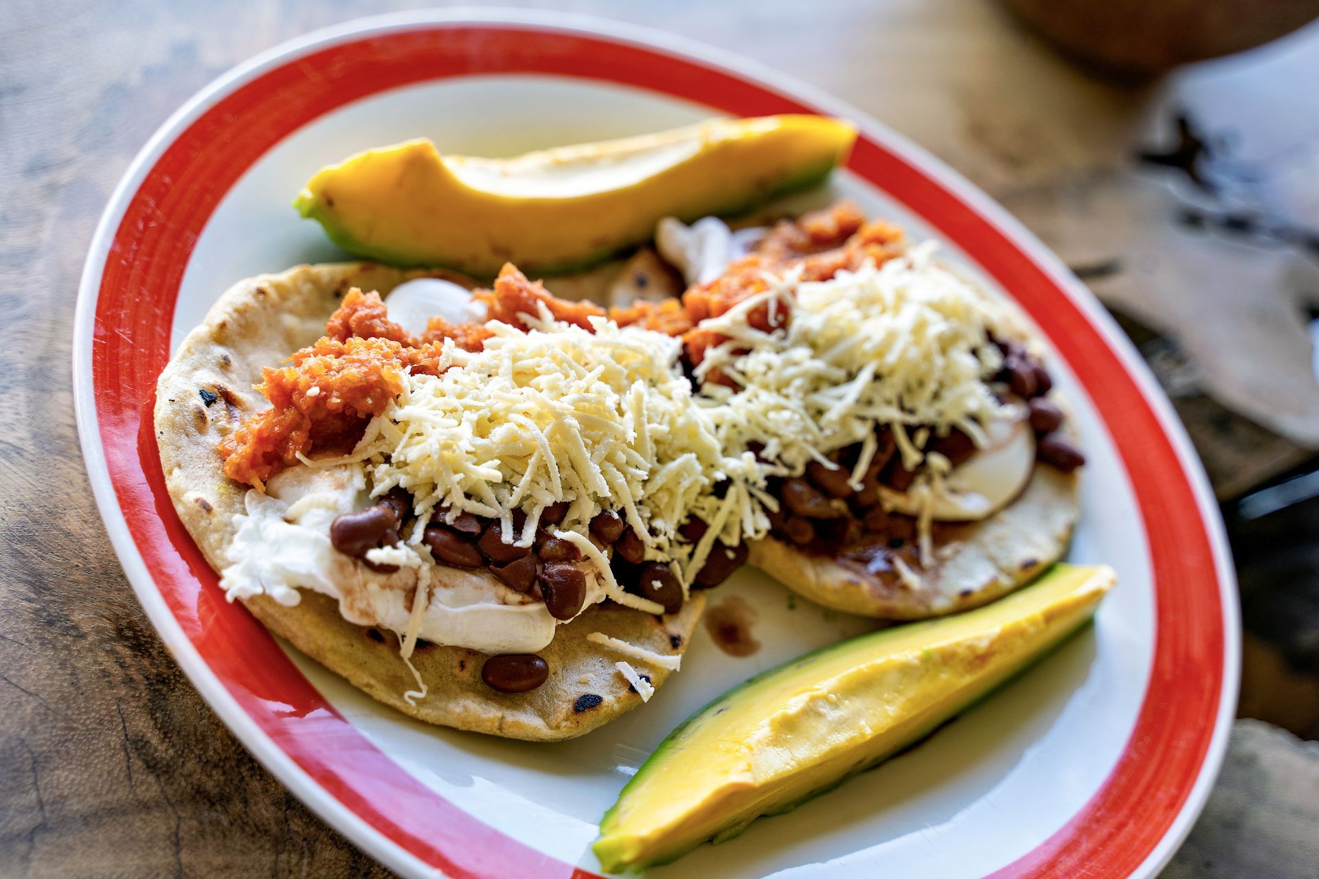 Two pupusas topped with beans, cheese, and sauce, served with avocado on a red-rimmed plate.