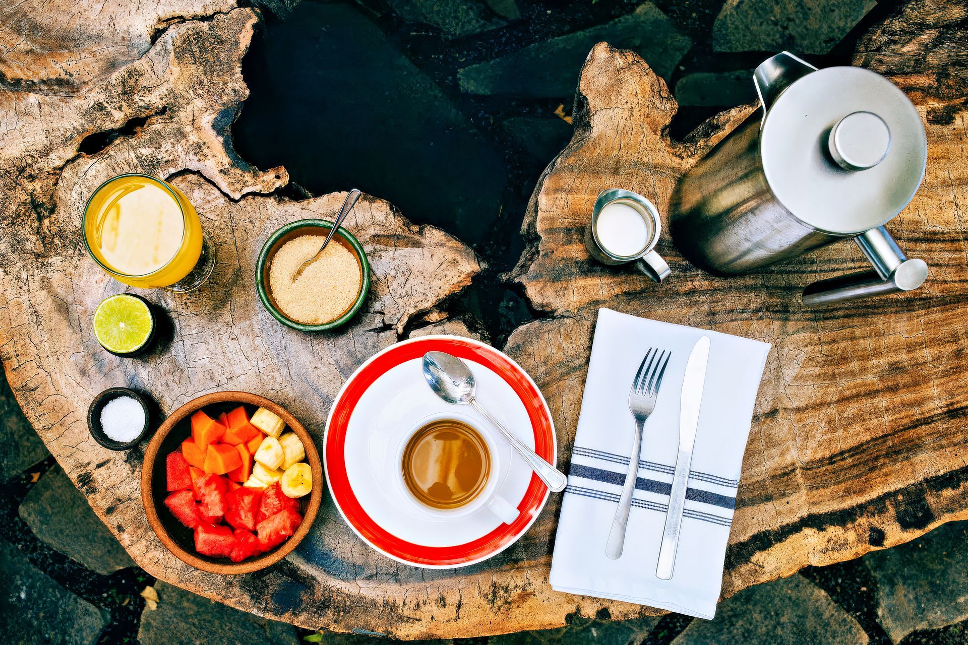 Breakfast spread on a rustic wooden table: juice, fruit, coffee, sugar, cream, cutlery.