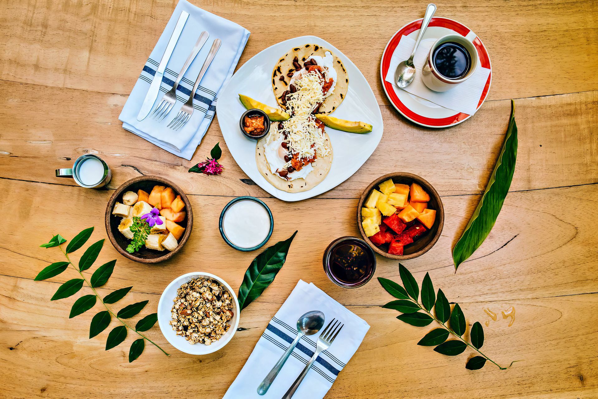 Breakfast spread on a wood table includes fruit bowls, pupusas, coffee, and utensils.