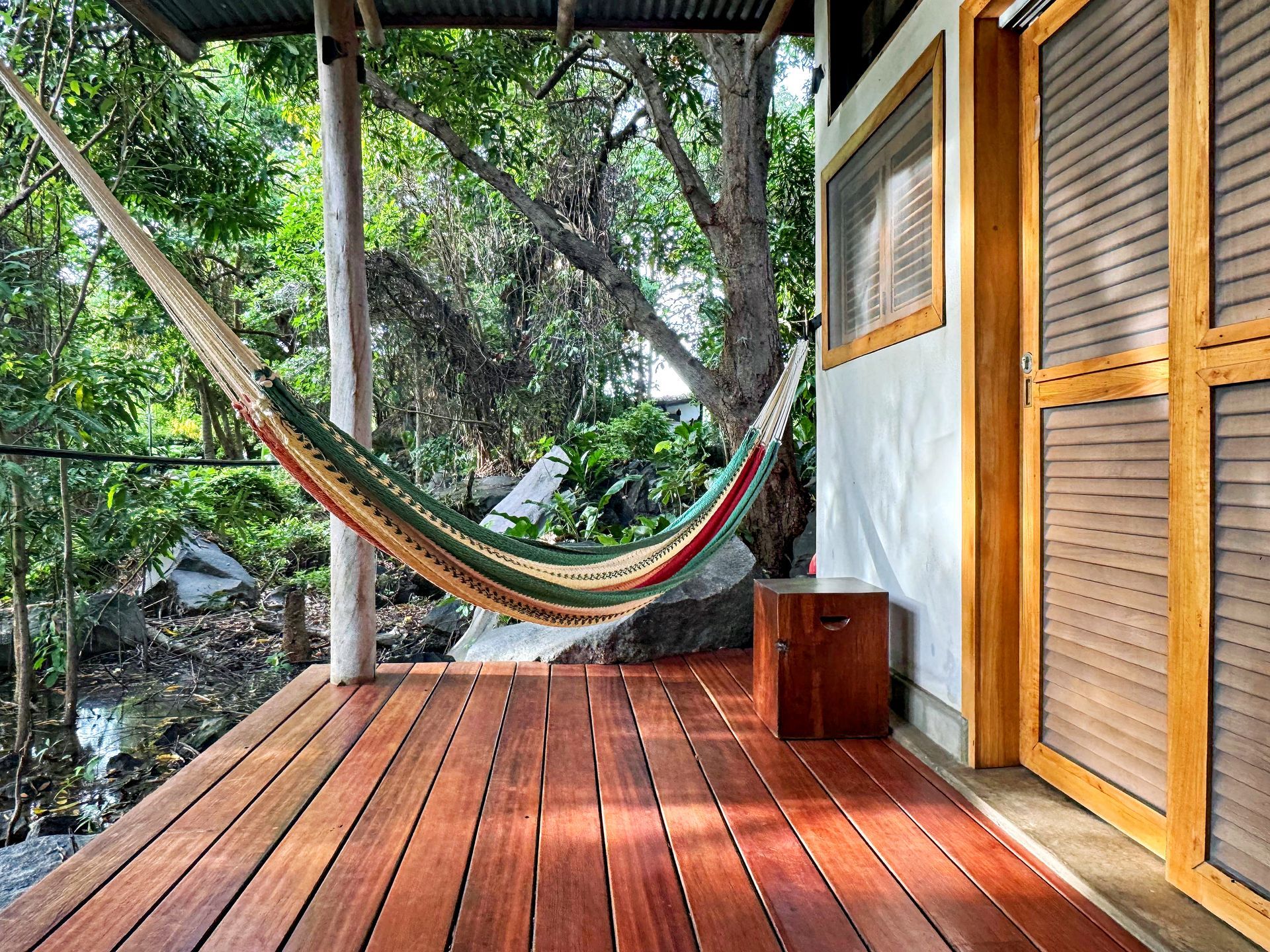 Wooden deck with a hammock, looking onto lush greenery and a cabin.