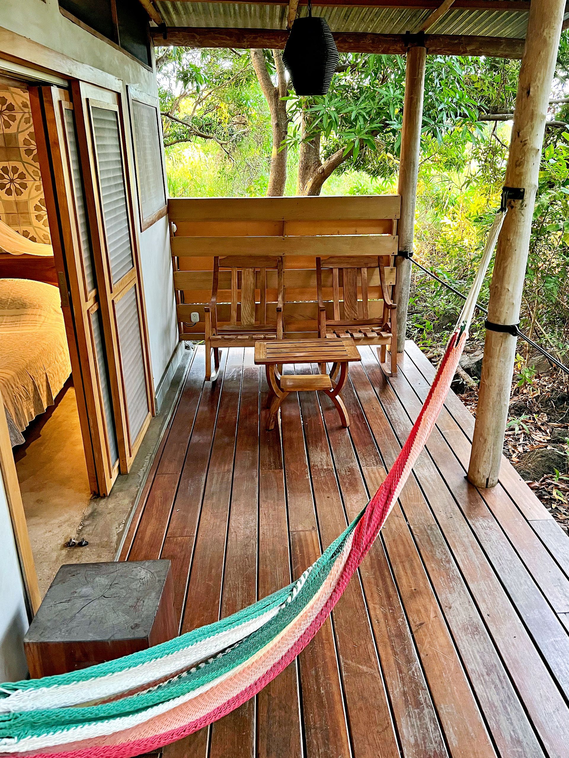 Wooden cabin porch with hammock, bench, and small table; surrounded by trees.