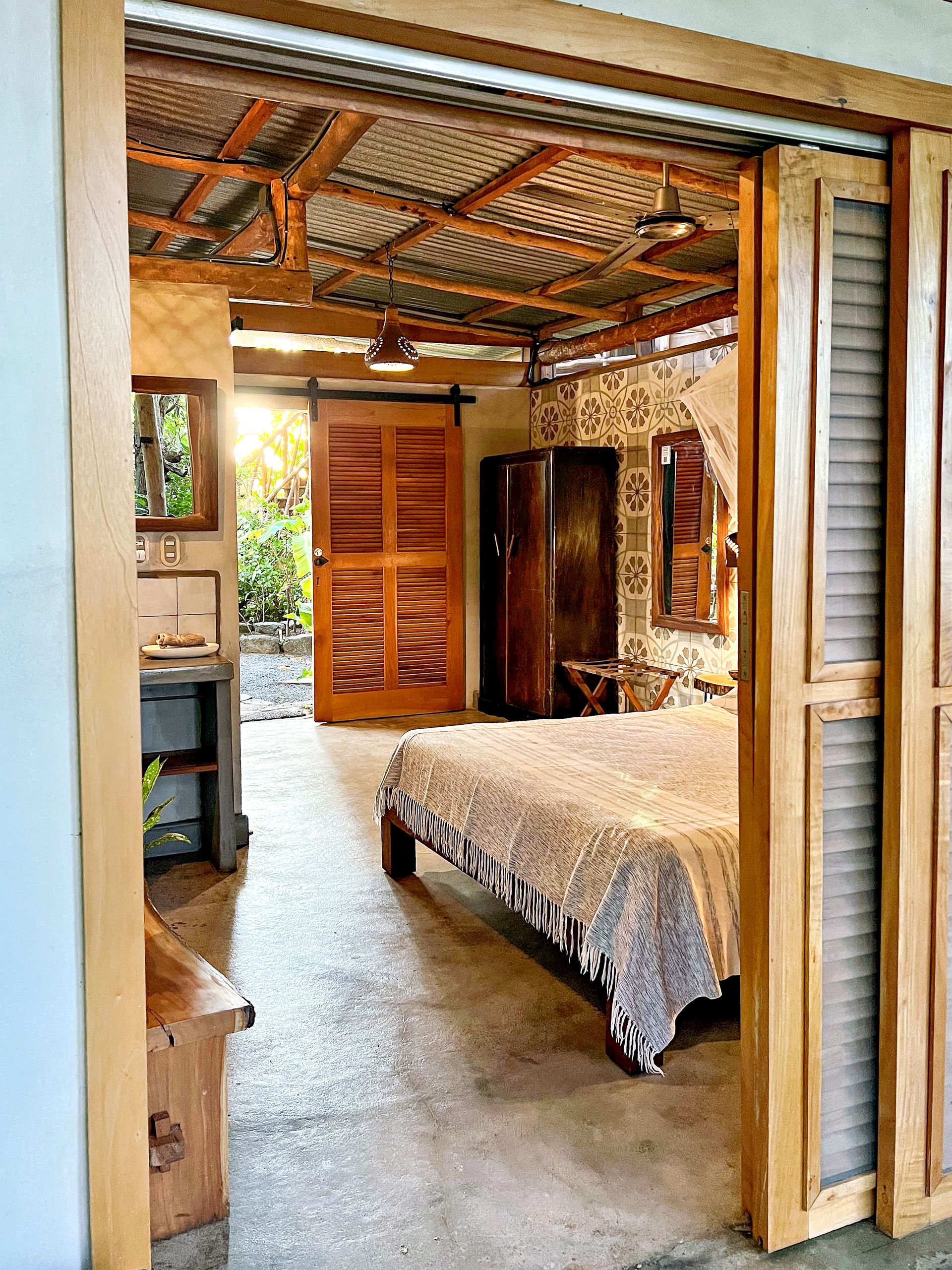 Bedroom interior: wooden doors open to a bed, dresser, and natural light from an outdoor view.