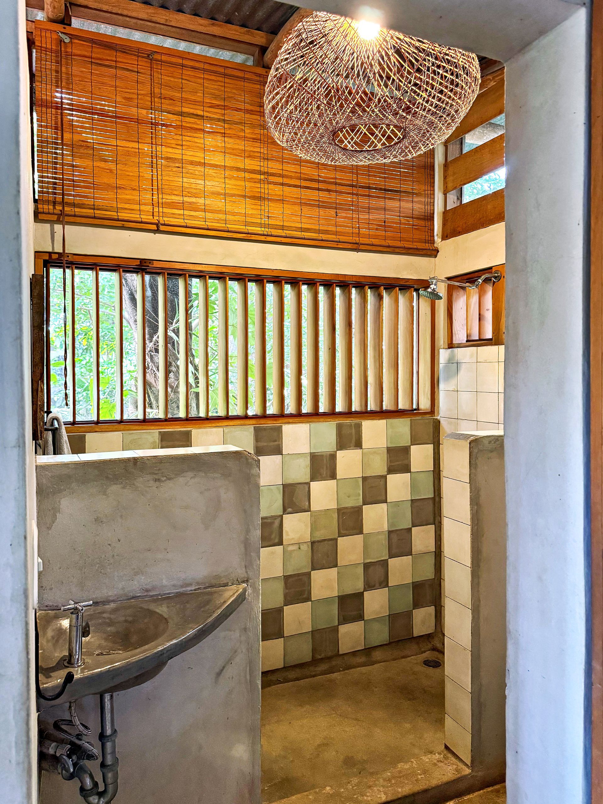 Rustic bathroom with checkerboard tiles, bamboo blinds, concrete sink, and woven light fixture.