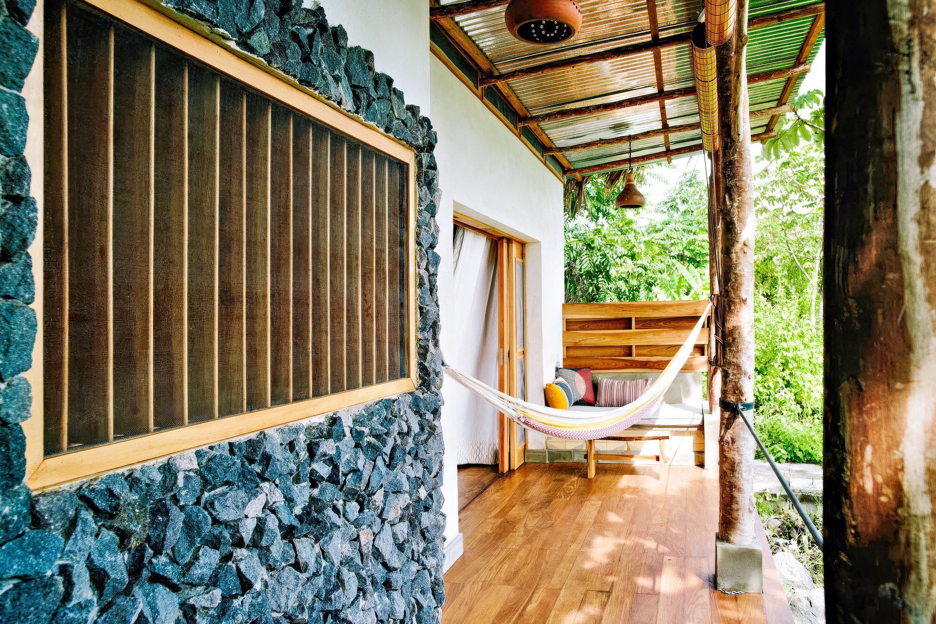 Veranda with hammock; stone wall with window; corrugated metal roof; lush green foliage in background.