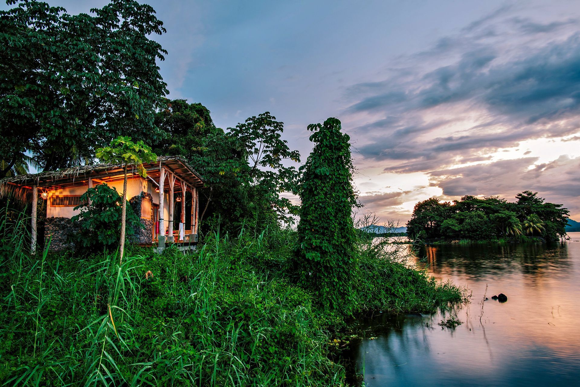 Lush green shoreline with a rustic building at dusk. Island, water, and cloudy sky in the distance.