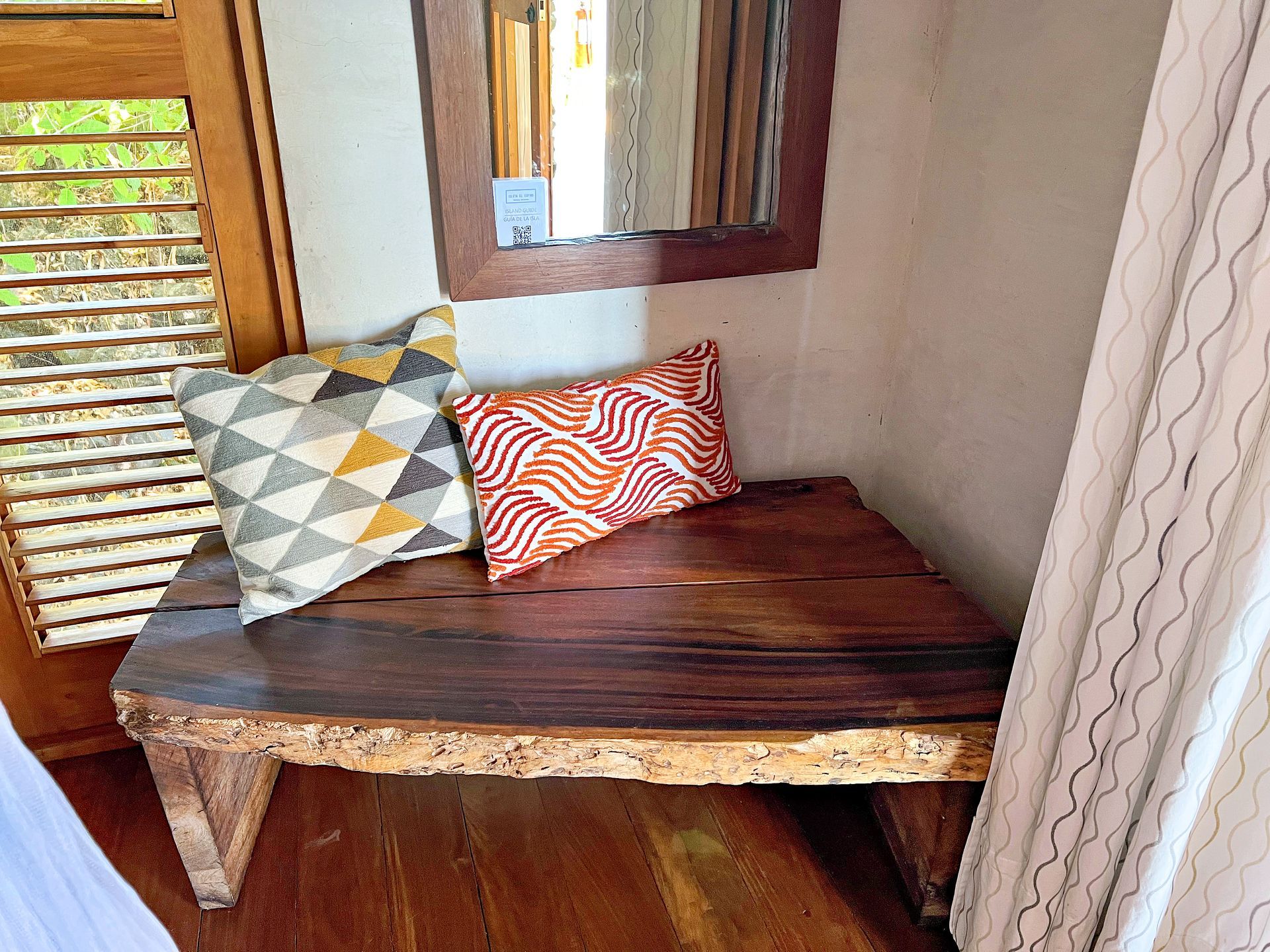 Wooden bench with two patterned pillows, under a mirror. Room corner with wooden blinds.