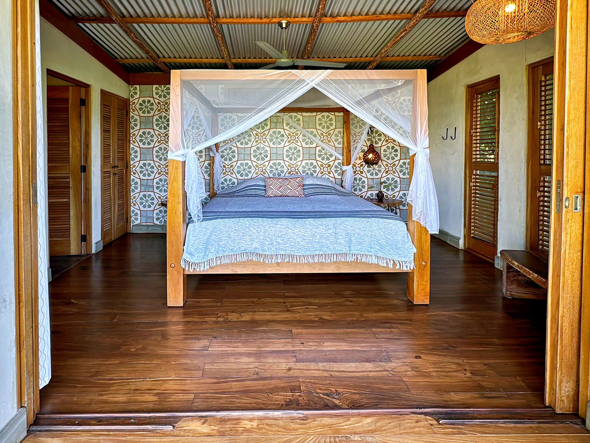 Wooden framed bed with mosquito net, in a room with open doors, wooden floors, and patterned wallpaper.