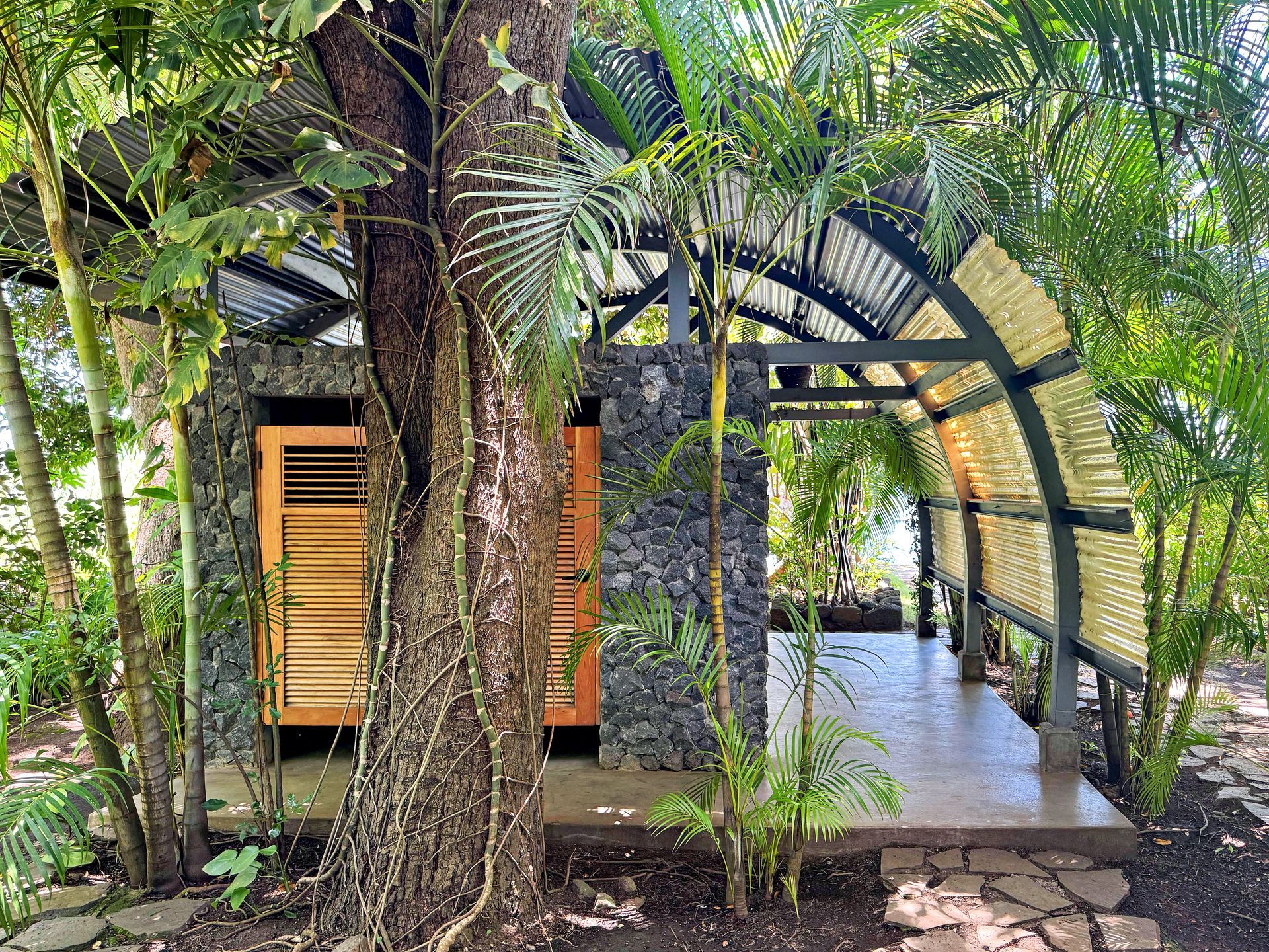Stone outdoor shower with a curved roof, surrounded by lush green plants.