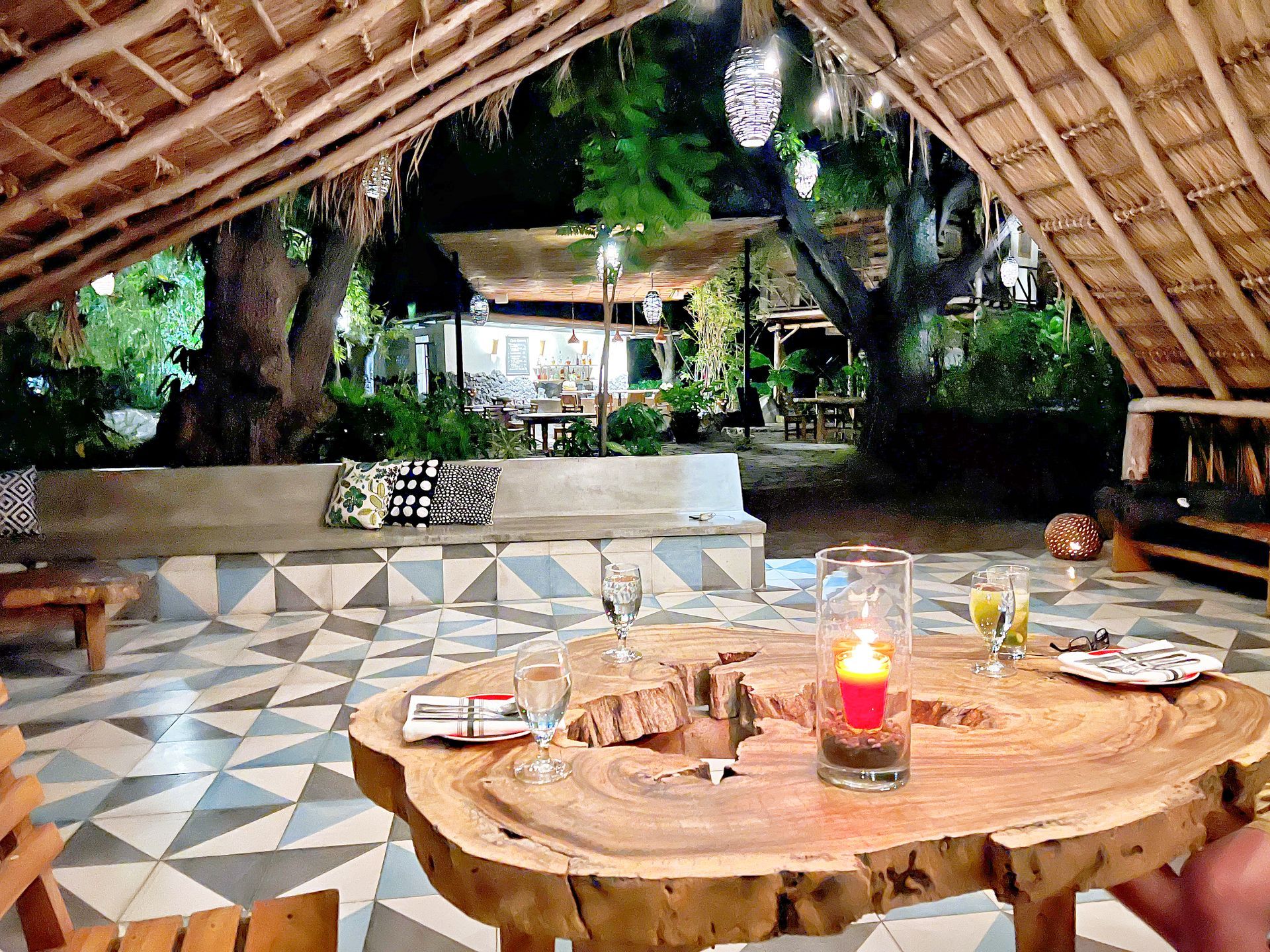 Outdoor lounge area at night, rustic wood table with drinks, lit candle, tiled floor, thatched roof.
