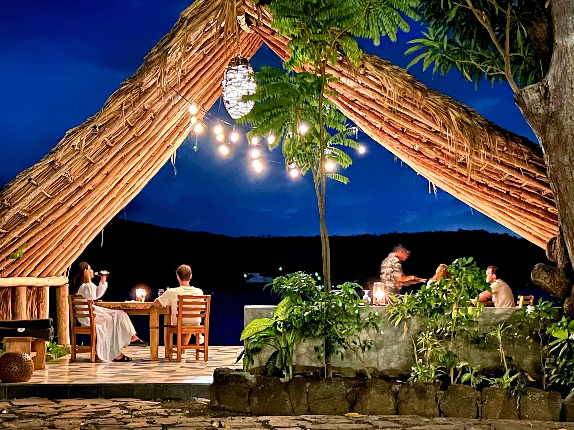 People dining at an open-air restaurant under a thatched roof, overlooking a dark landscape. Lit by string lights.