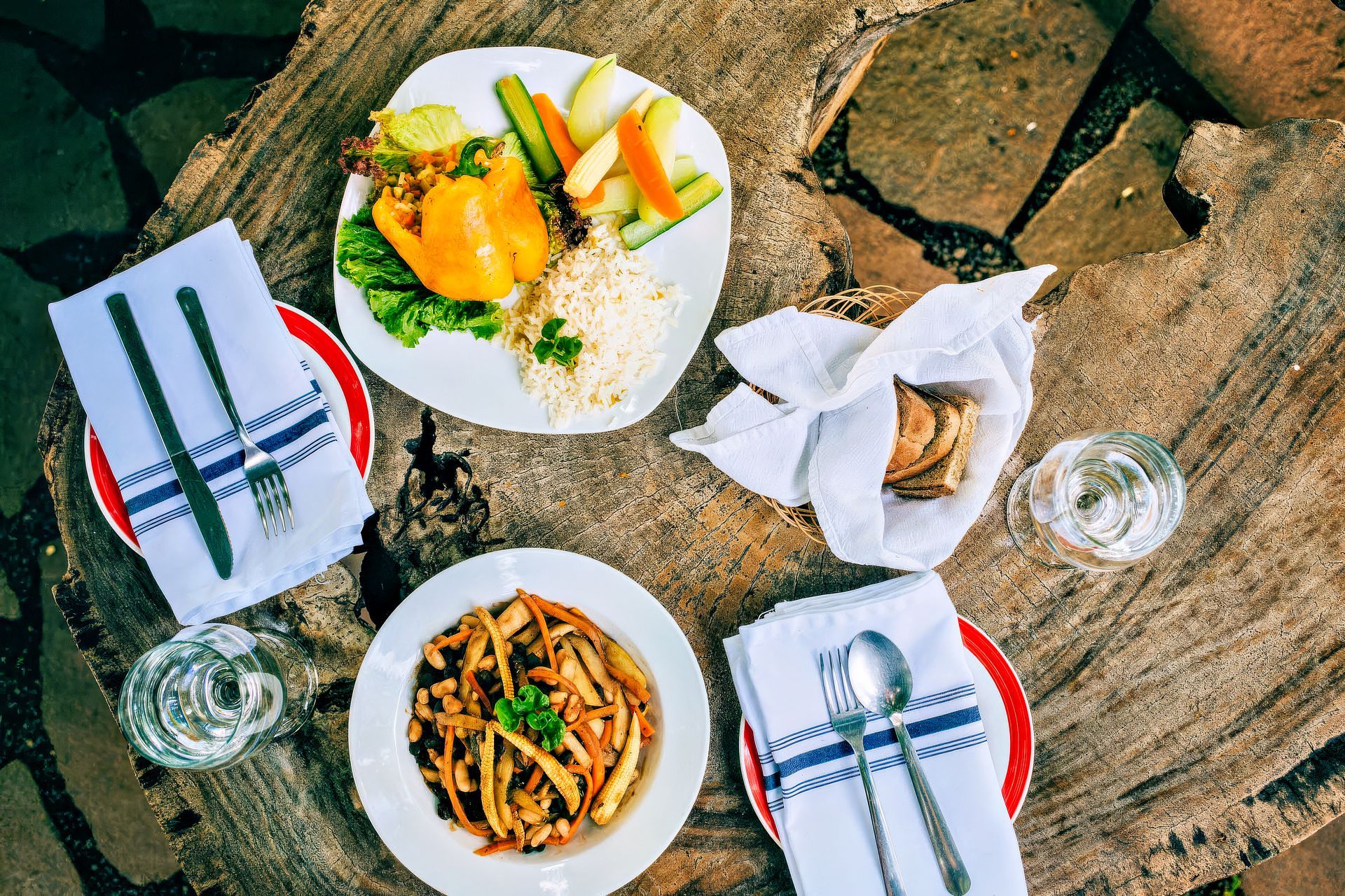 Overhead view: Two plates of food on wood table, with napkins, glasses, silverware.