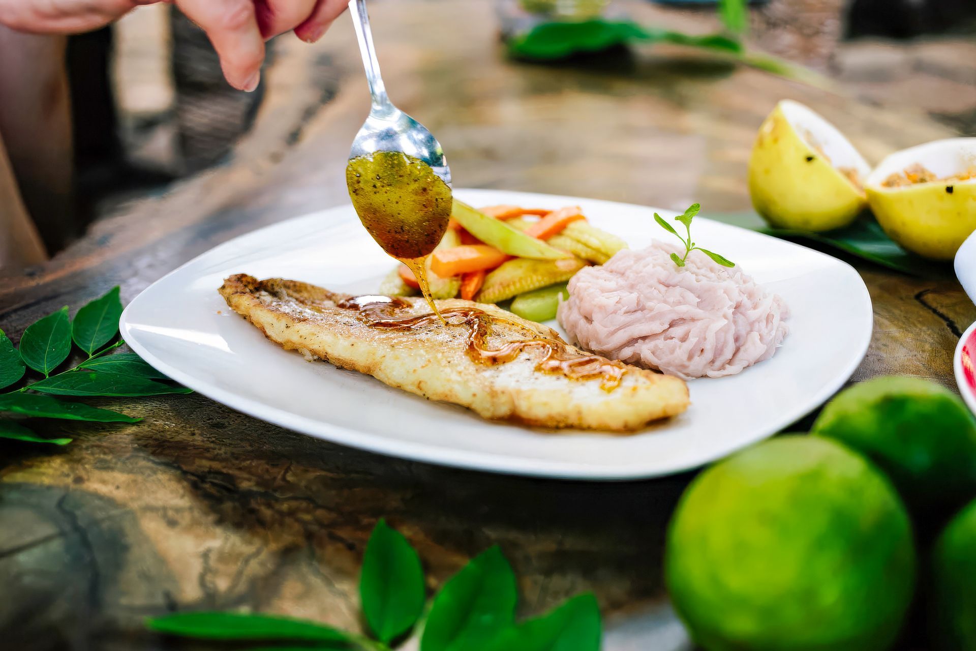 Person drizzling sauce over a grilled fish dish on a white plate.  Side of vegetables and mashed food