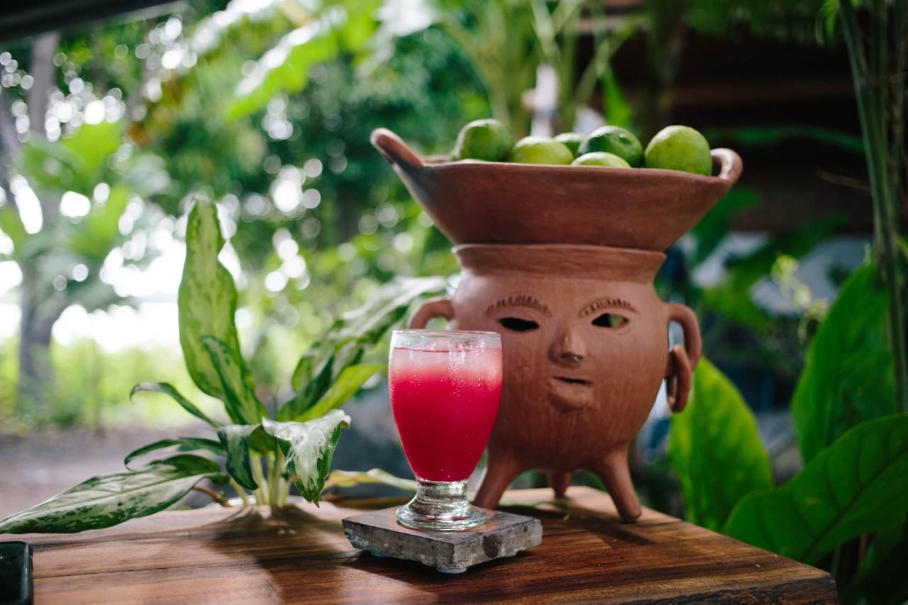 Red drink in a glass next to a terracotta pot with a face, and limes, on a wooden table.