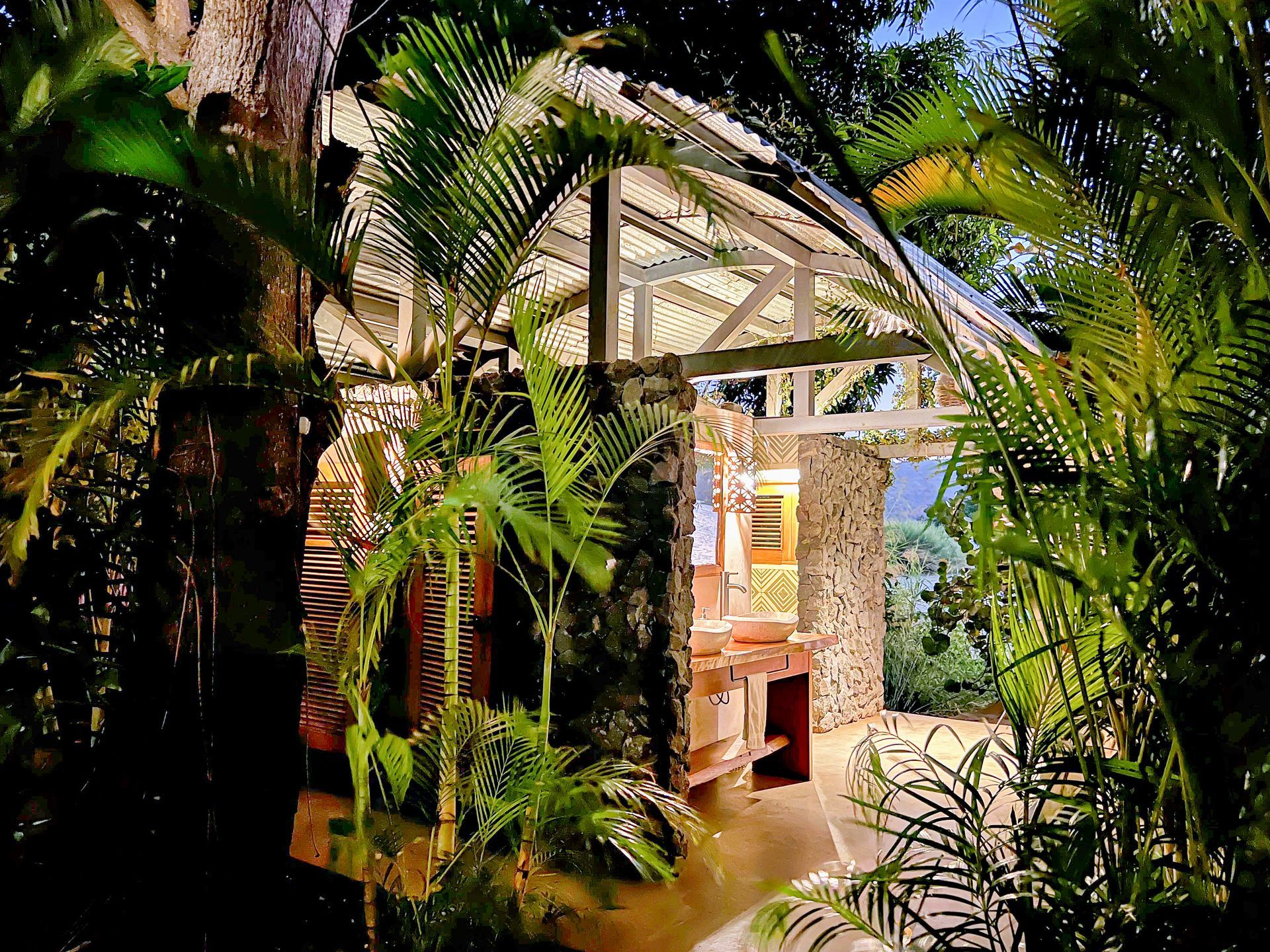 A secluded, open-air structure in lush greenery, featuring white beams and stone walls, lit at dusk.
