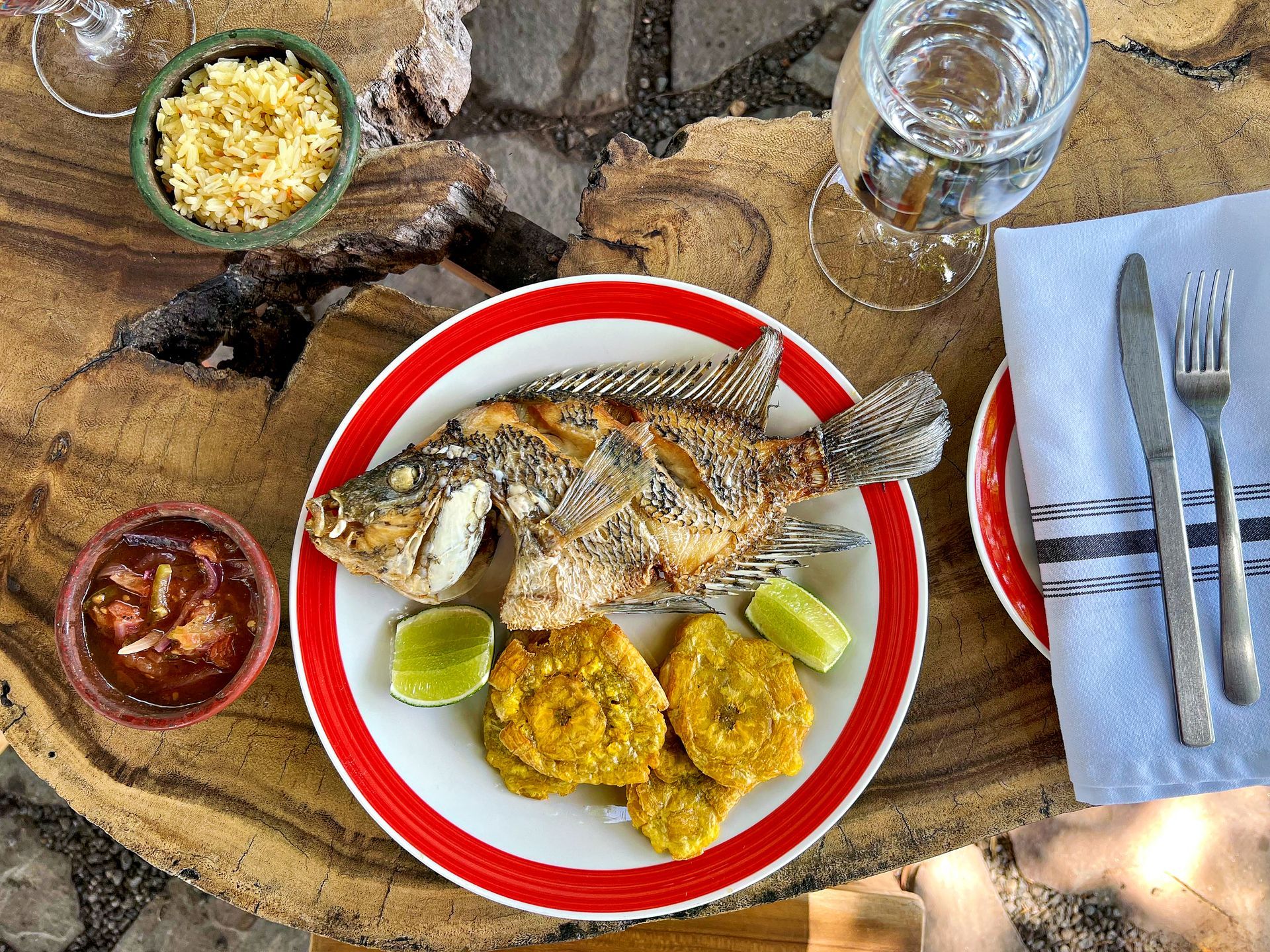 Fried fish with tostones, rice, and sauce on a wooden table, with lime wedges and a glass of water.