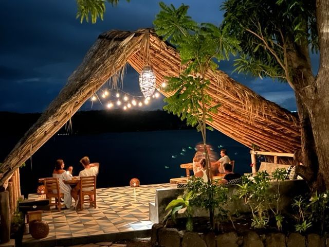 People dine outdoors under a thatched roof illuminated by string lights, overlooking a dark body of water.