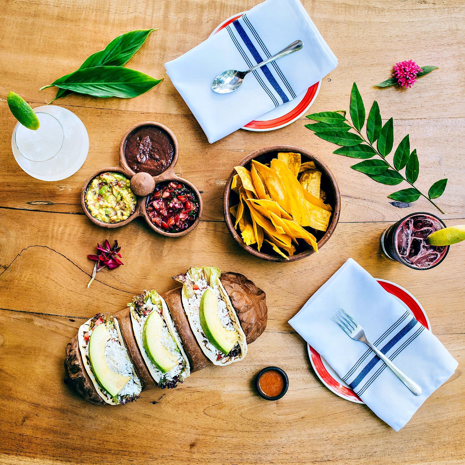 Wooden table with tacos, chips, dips, and drinks, a place setting on each side.