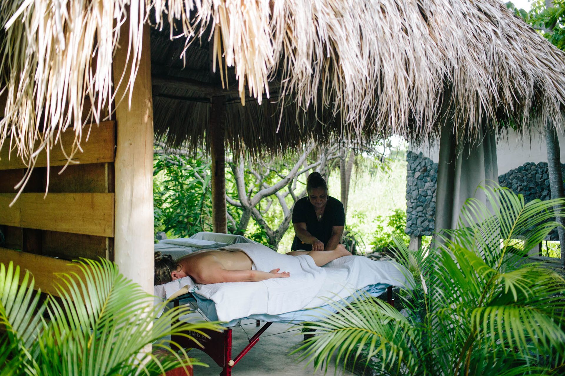 Massage outdoors under a thatched roof; therapist kneads patient's leg.