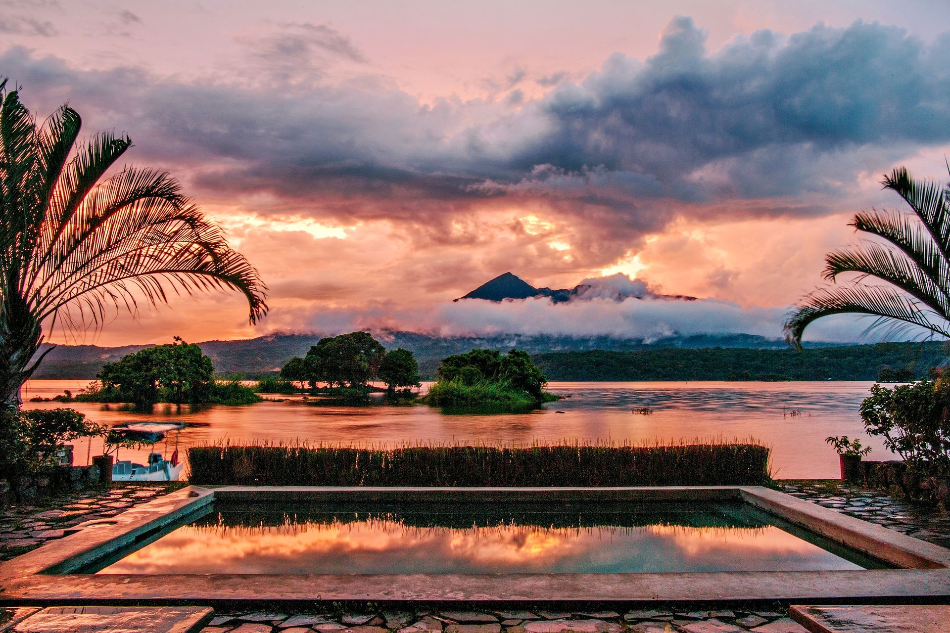 Sunset over lake with pool; pink and orange sky, palm trees, dark clouds.