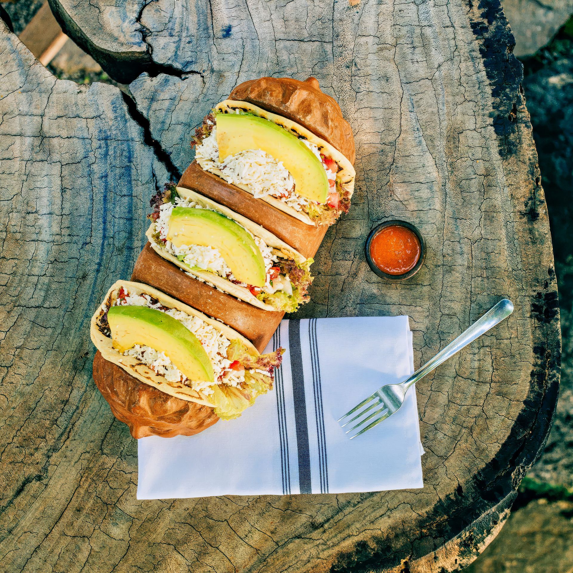 Tacos with avocado slices on a wooden stump. Small sauce dish, fork and napkin present.