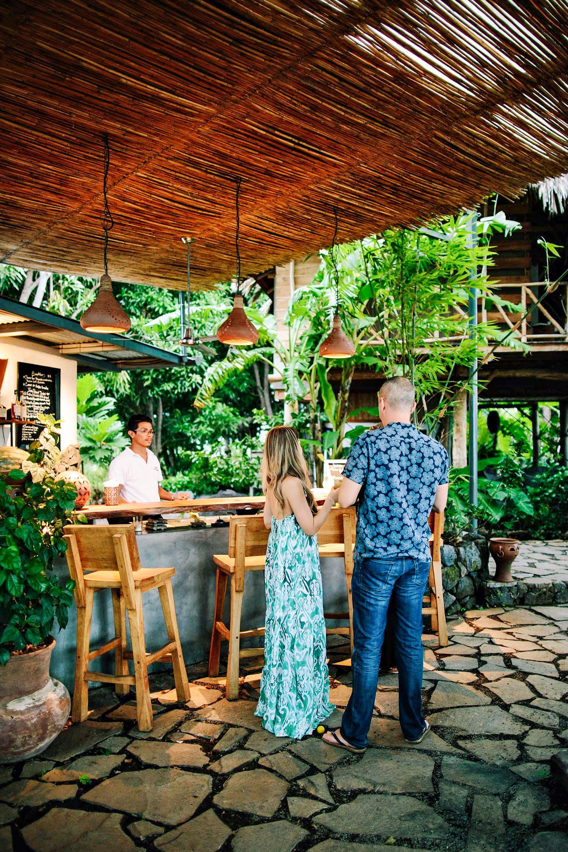 Couple ordering drinks at outdoor bar; bartender behind counter. Lush greenery and straw roof.