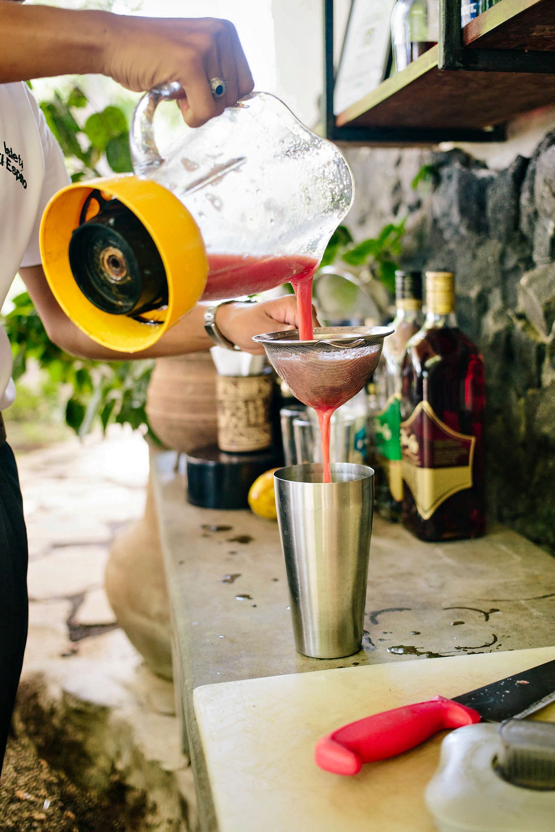 Bartender strains a red liquid into a metal shaker outdoors; bottles and stone wall in the background.