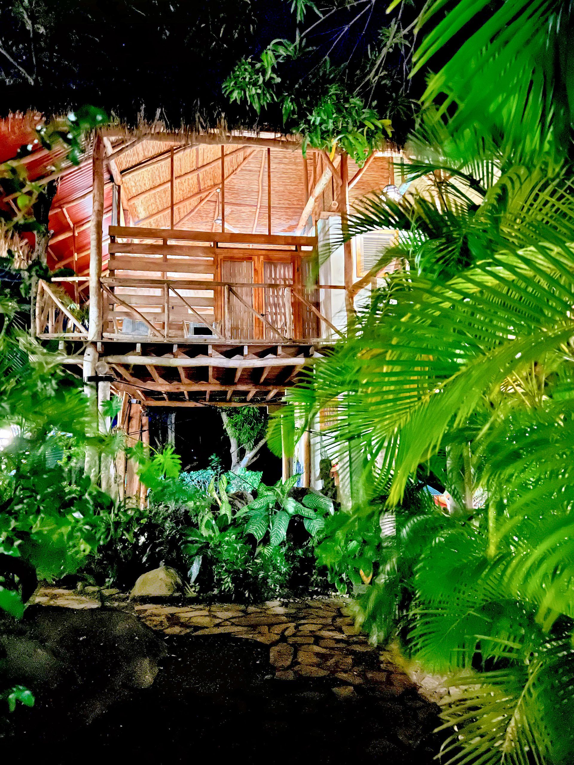 Wooden treehouse at night, surrounded by lush green foliage and lit path.