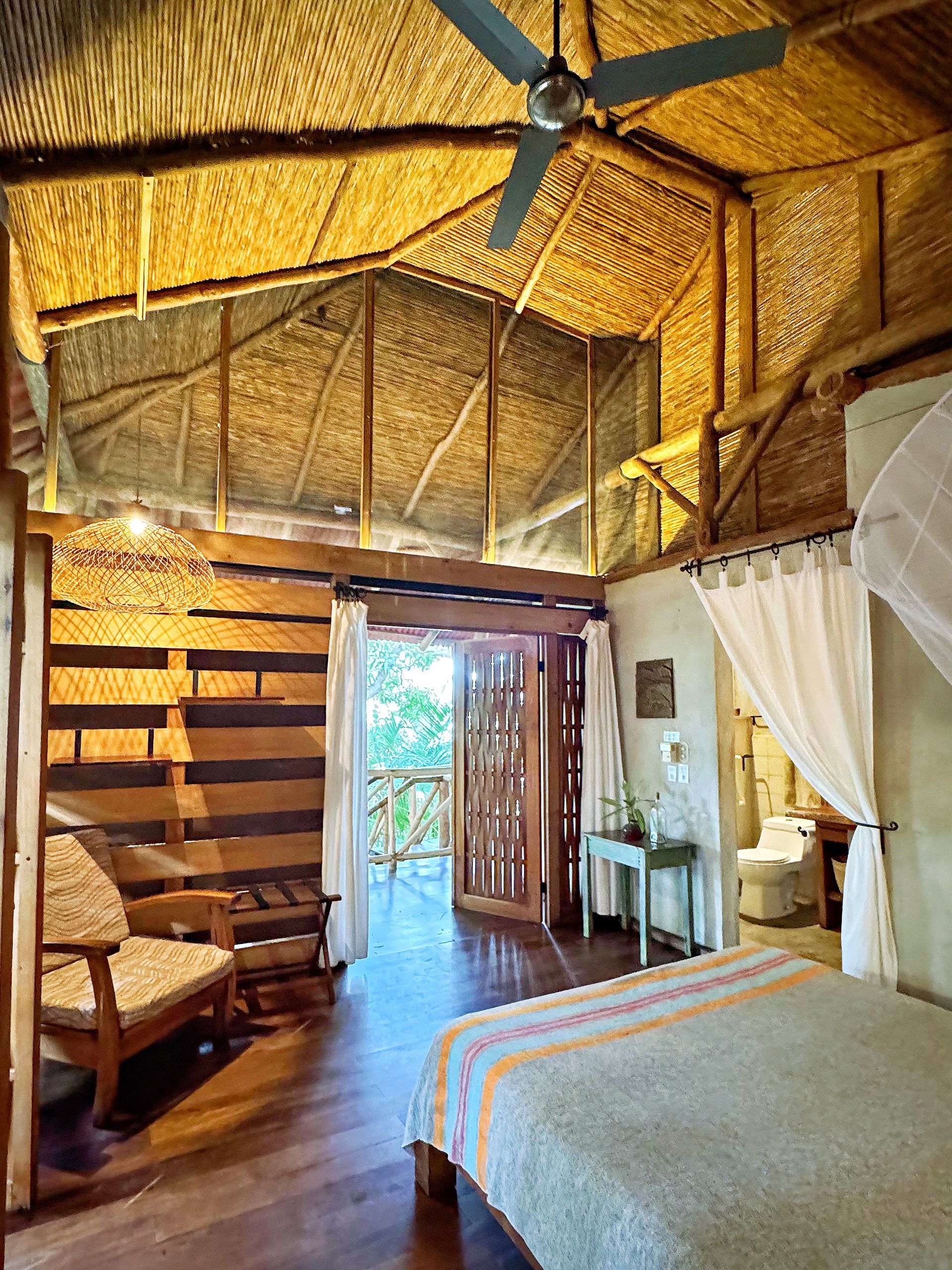 Wooden cabin interior with bed, seating, balcony view, and thatched roof.