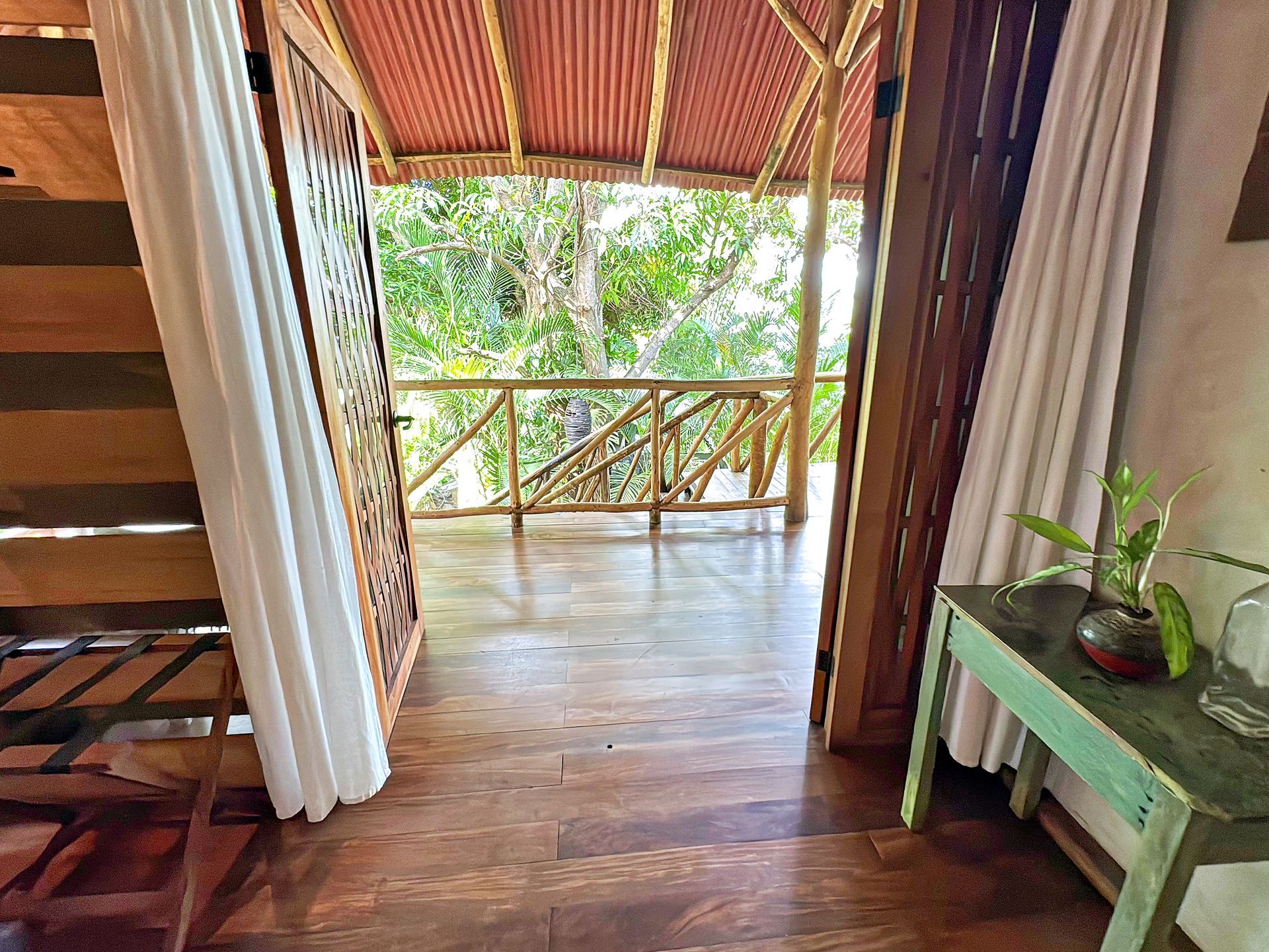 Wooden-floored room opens to a balcony with a jungle view; wooden stairs on the left, a green table with plants on the right.