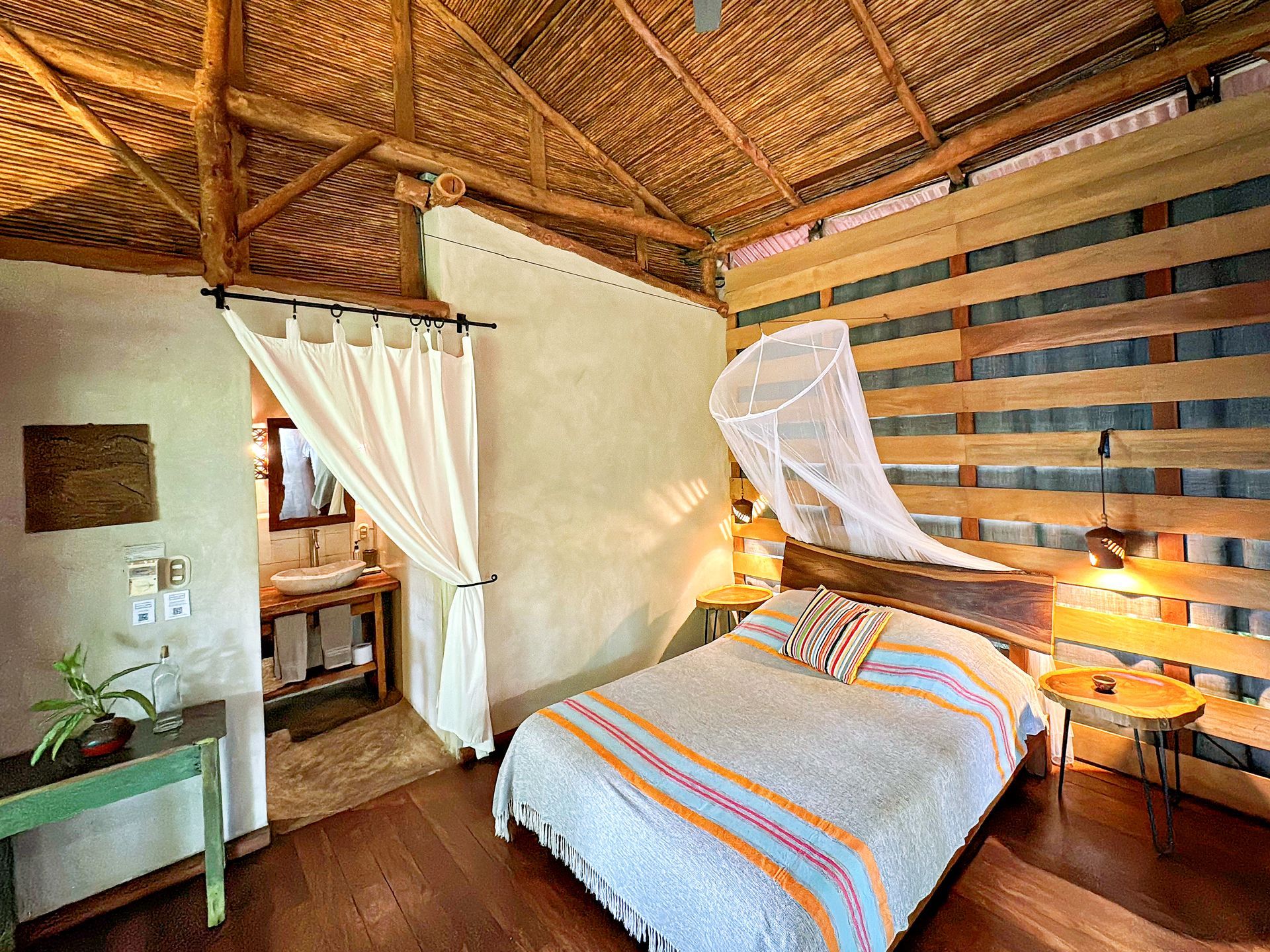Bedroom with a wooden bed, mosquito net, and woven wooden walls. Bathroom with sink visible.