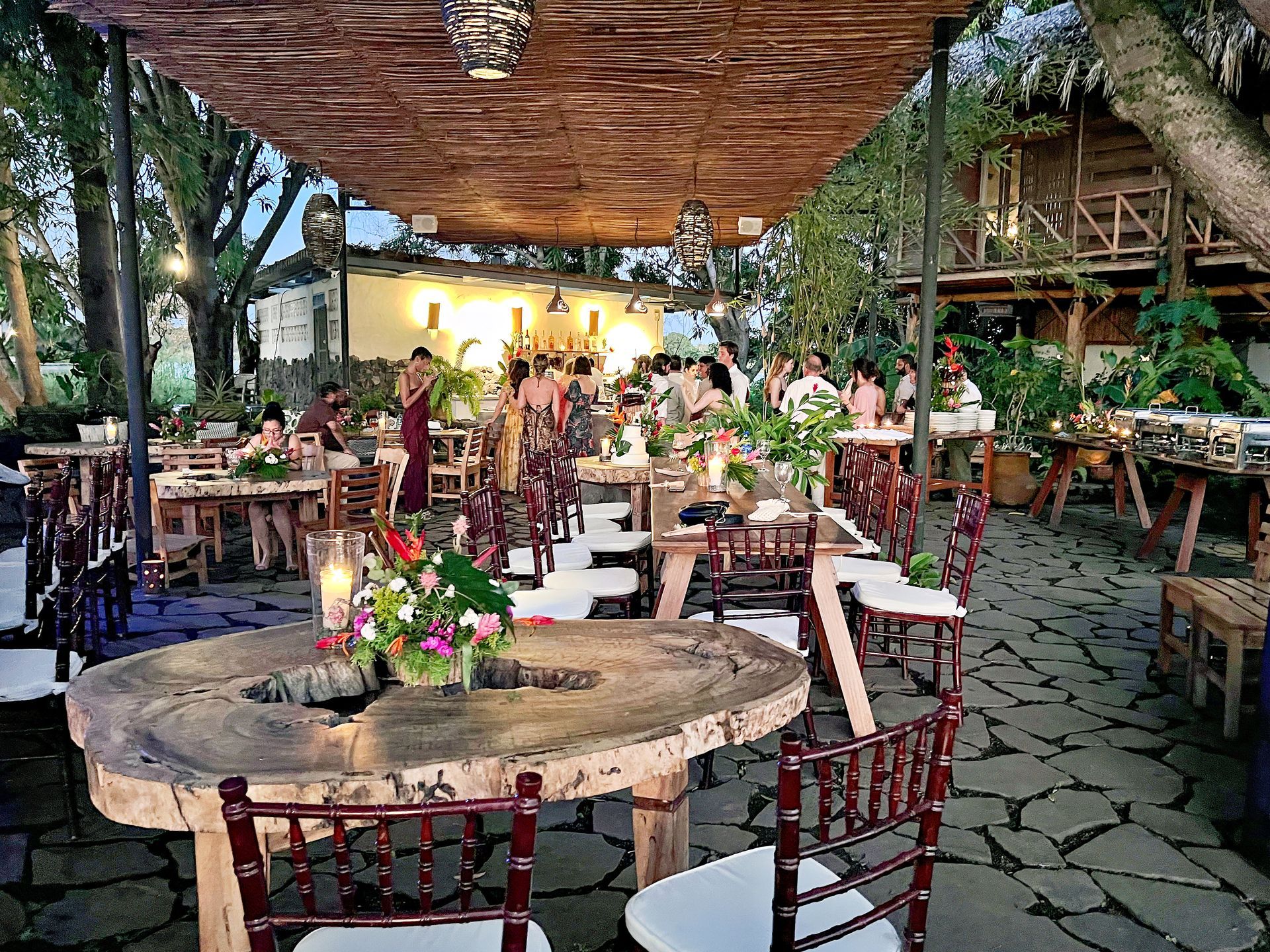 Outdoor wedding reception with wooden tables, floral centerpieces, and guests gathered under a rustic canopy.