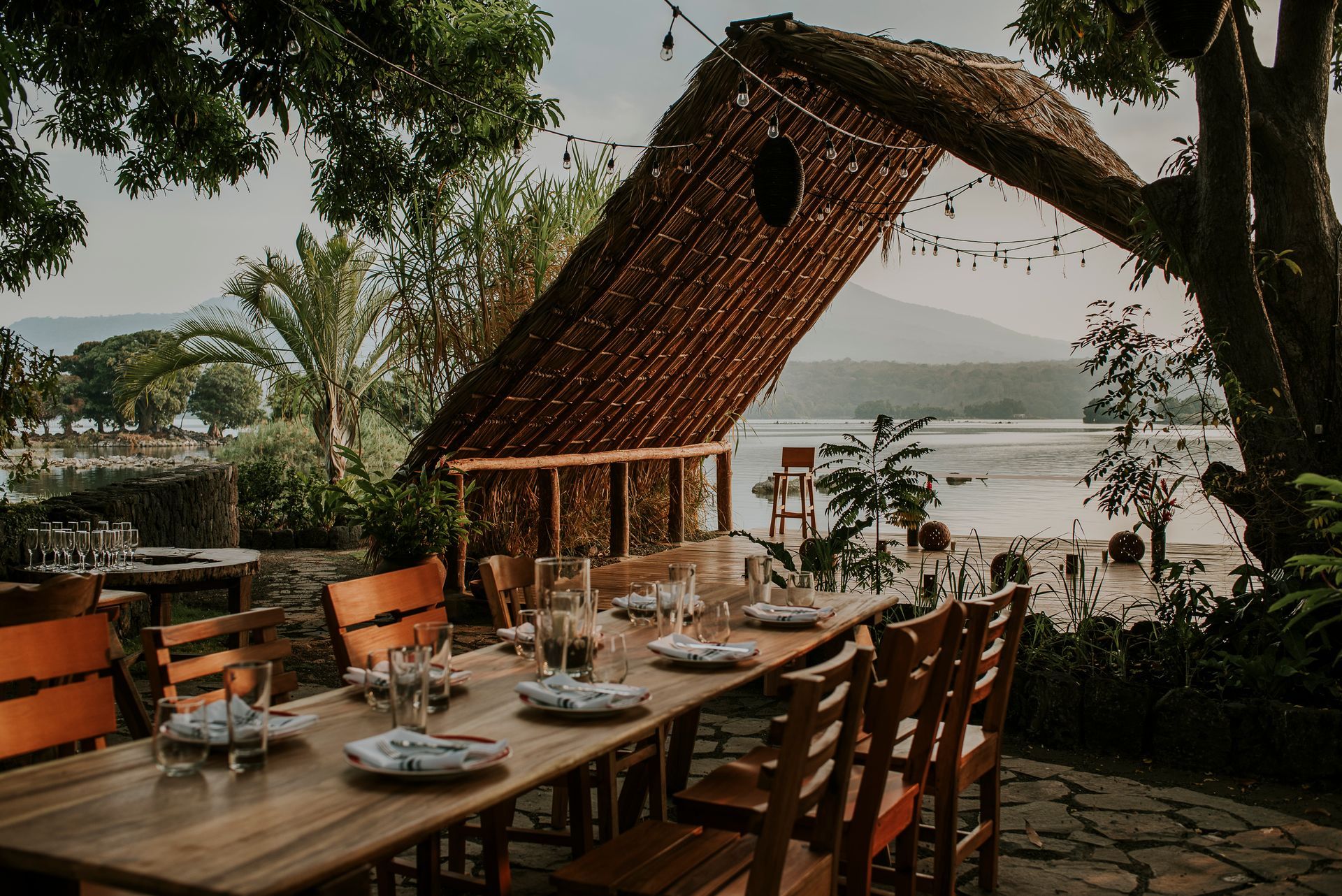 Outdoor dining table set for a meal, with a water view, under a thatched roof.