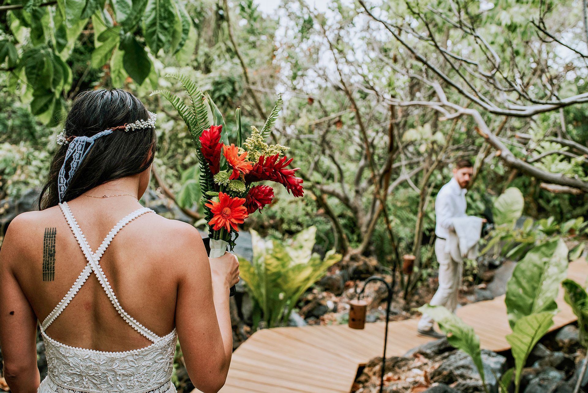 Bride in a white dress carrying flowers, facing away, walks towards the groom on a wooden path in a lush, green environment.