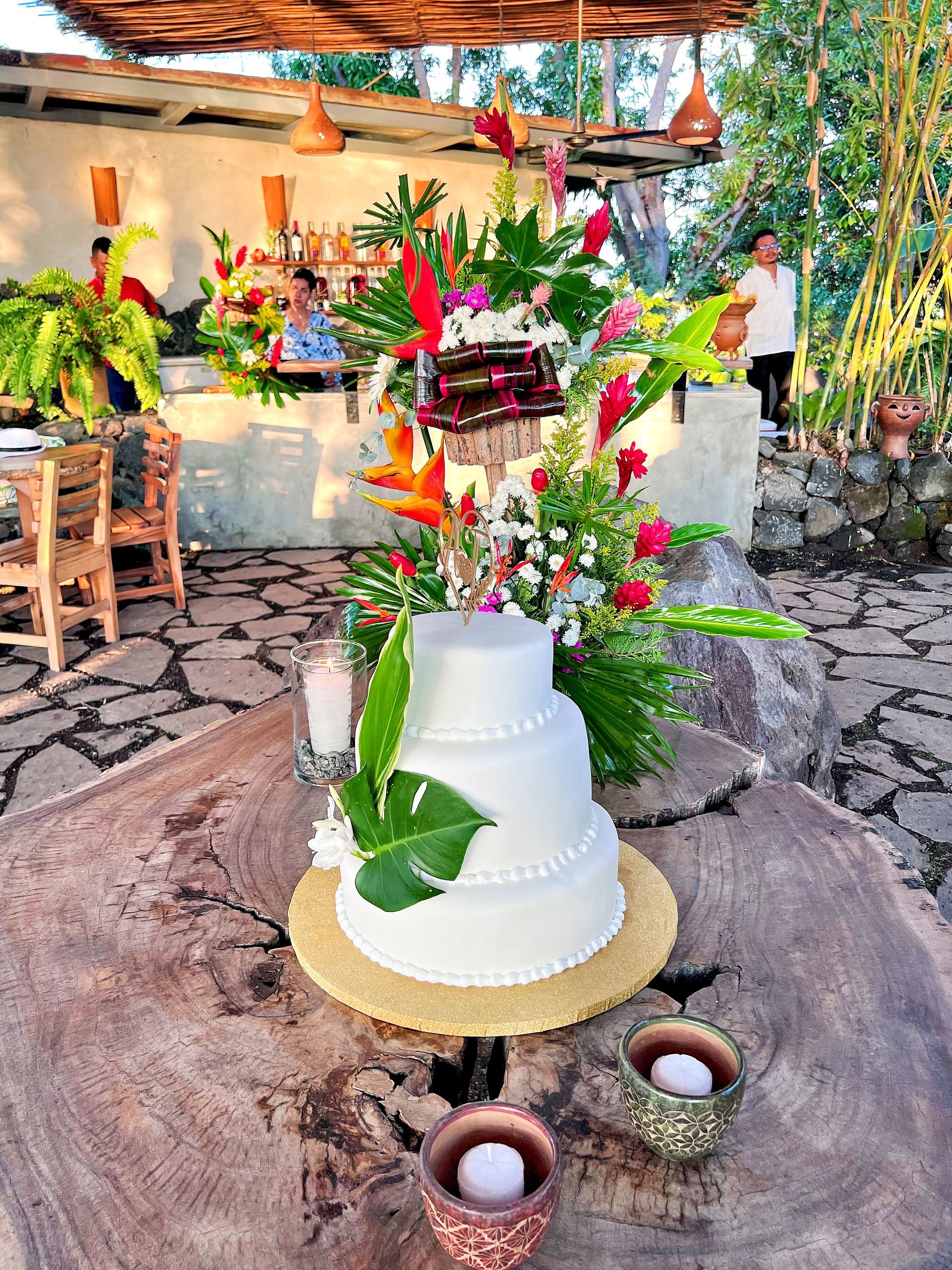 Wedding cake on a rustic wooden table, decorated with tropical flowers. Outdoor reception setting.