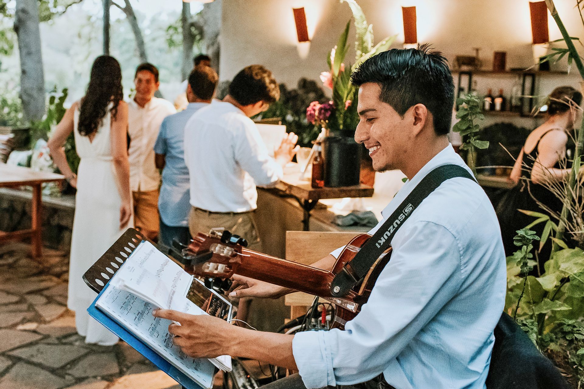 Man playing guitar, reading music sheets, smiling at an outdoor event. Guests in the background, warm lighting.
