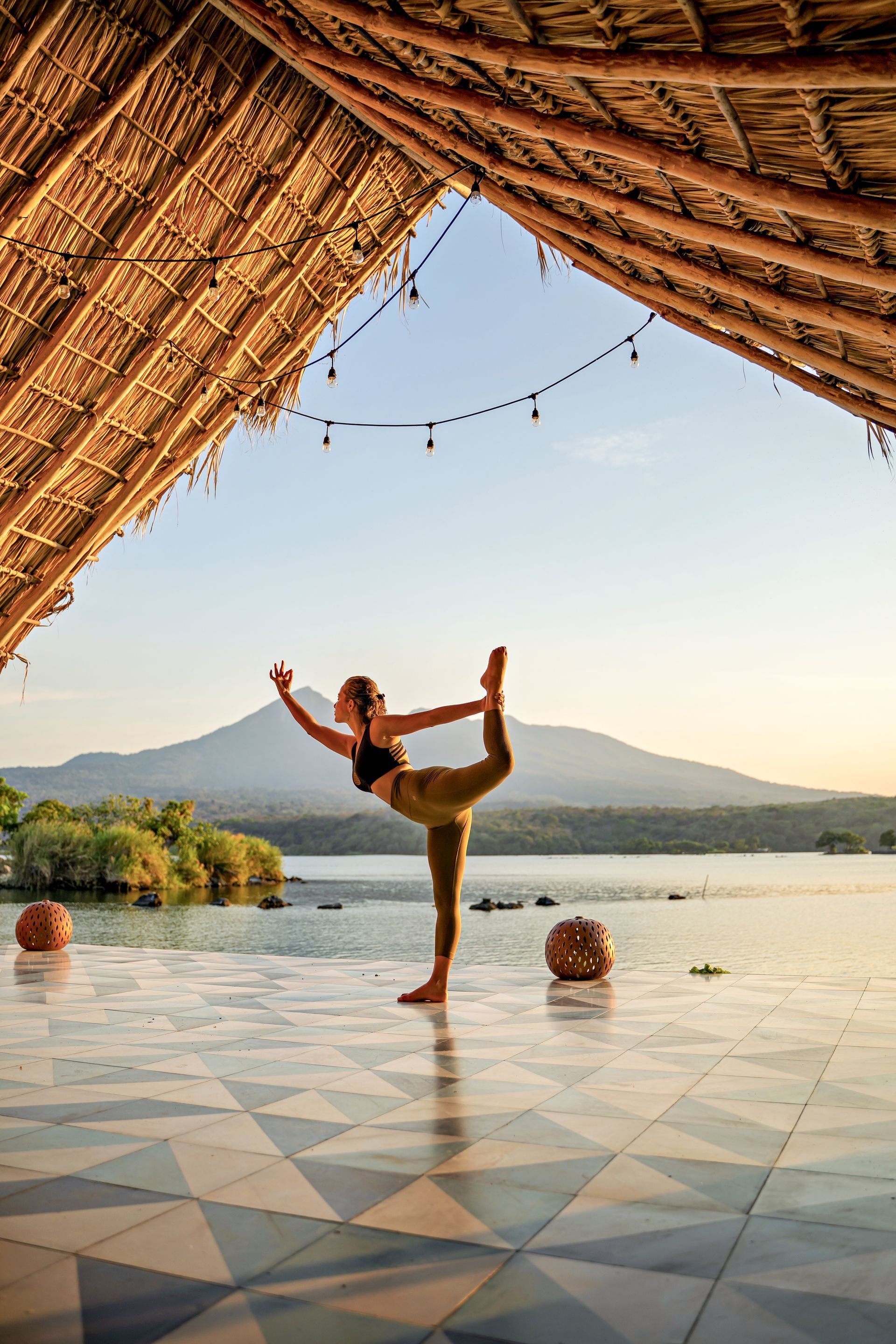Woman balances in yoga pose on a tiled platform, facing a lake and mountain. Under a thatched roof, sunlight glows.