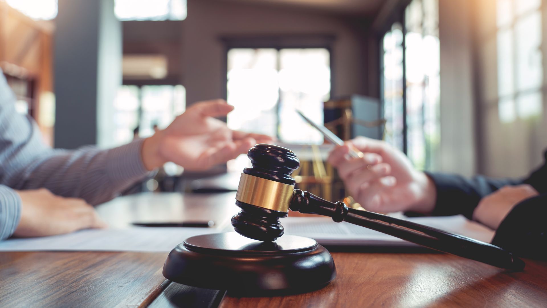 A judge's gavel rests on a wooden table in front of two people, one gesturing while speaking during a legal consultation. A judge's gavel rests on a wooden table in front of two people, one gesturing while speaking during a legal consultation.