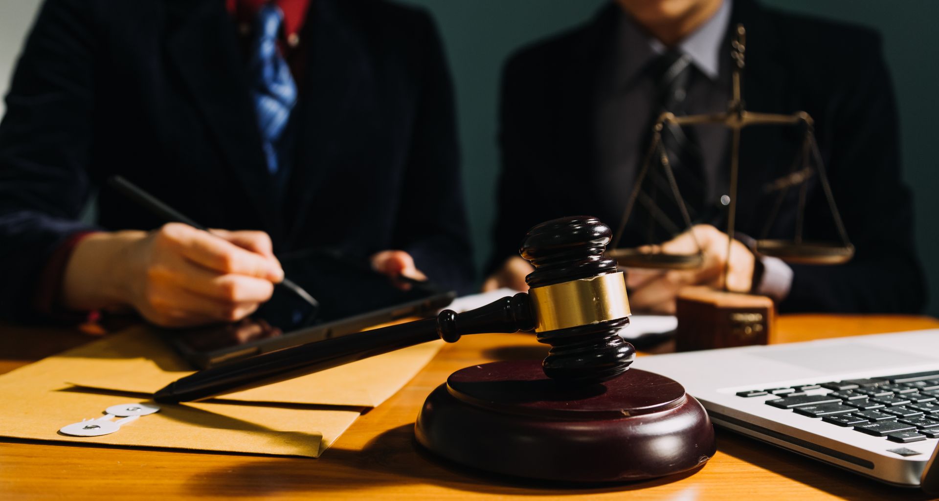 A judge's wooden gavel and a gold-accented scale of justice on a desk with two professionals working in the background.