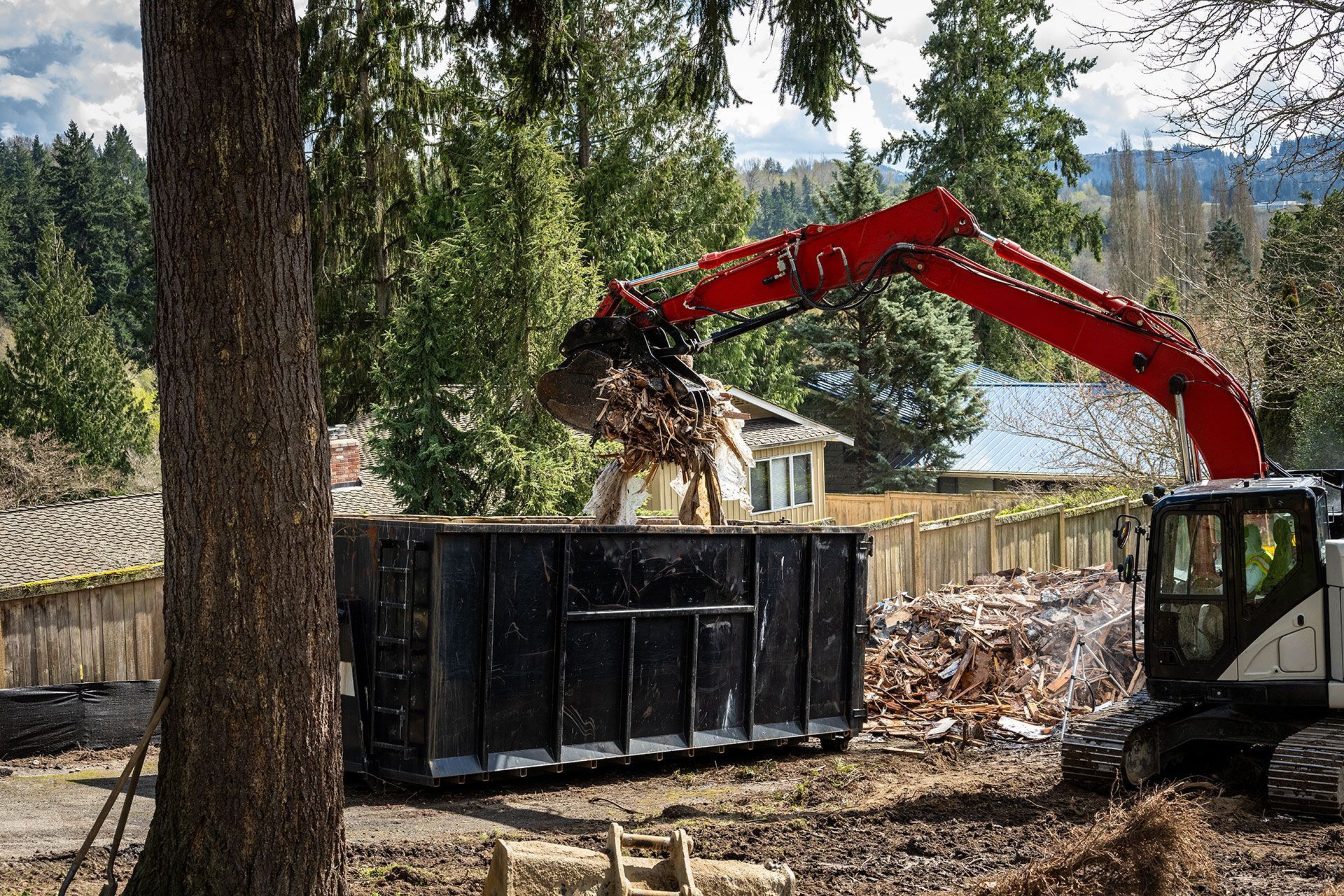 A red excavator loads debris into a black dumpster during a building demolition in a residential area.