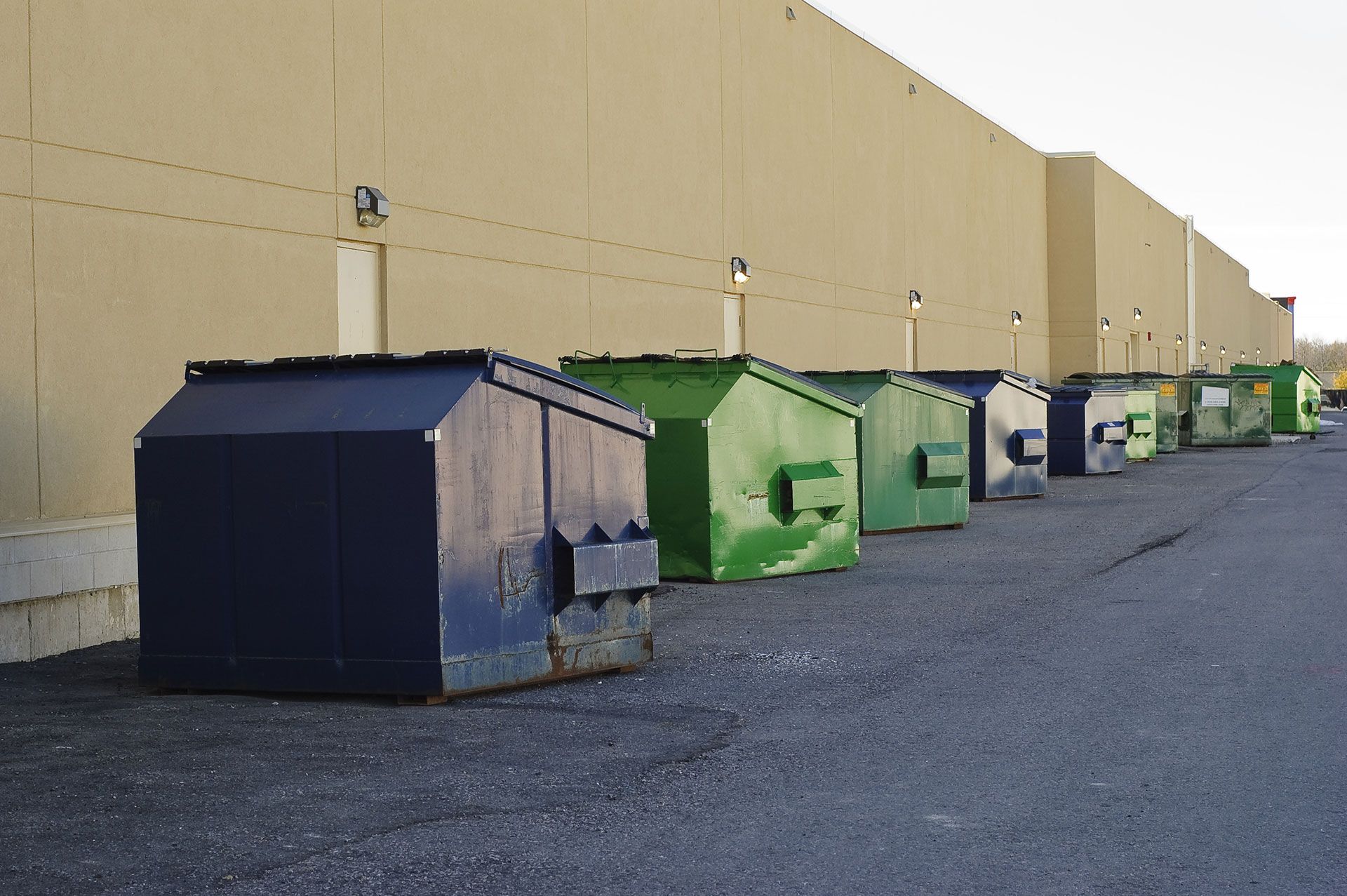 Row of dumpsters in various colors alongside a beige building; setting is outdoors.
