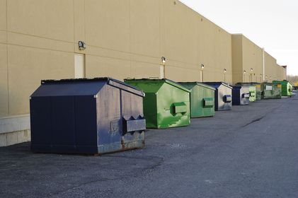 Row of dumpsters in various colors alongside a beige building; setting is outdoors.